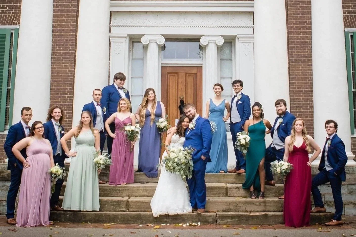 A large wedding party standing on the steps of a church with a wooden door. The bride and groom are in the center, with bridesmaids and groomsmen surrounding them, all dressed in formal attire. The women are wearing colorful dresses and holding bouquets, while the men are in blue suits with bow ties.