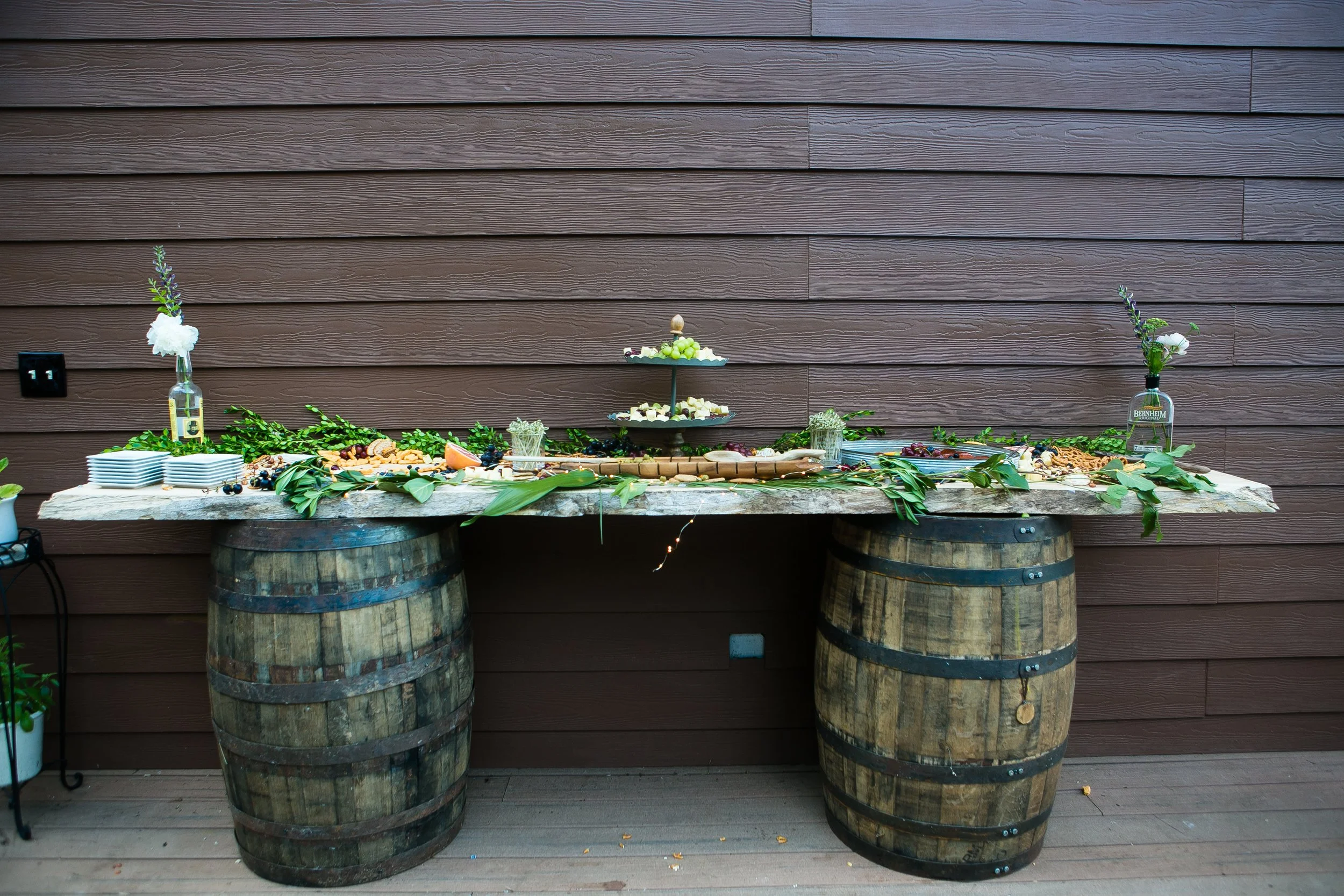 Buffet table set up outdoors on a wooden deck, supported by two large wine barrels. The table is decorated with green leaves and features plates of food, a tiered tray of desserts, and two glass bottles with flowers. The background is a wooden wall.