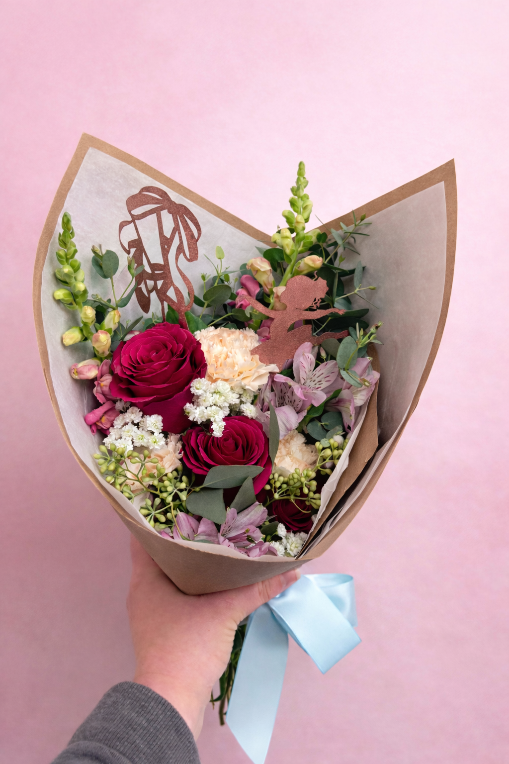 A hand holding a bouquet of various flowers, including pink roses, white carnations, and other mixed flowers, wrapped in brown paper with a light blue ribbon, set against a pink background.