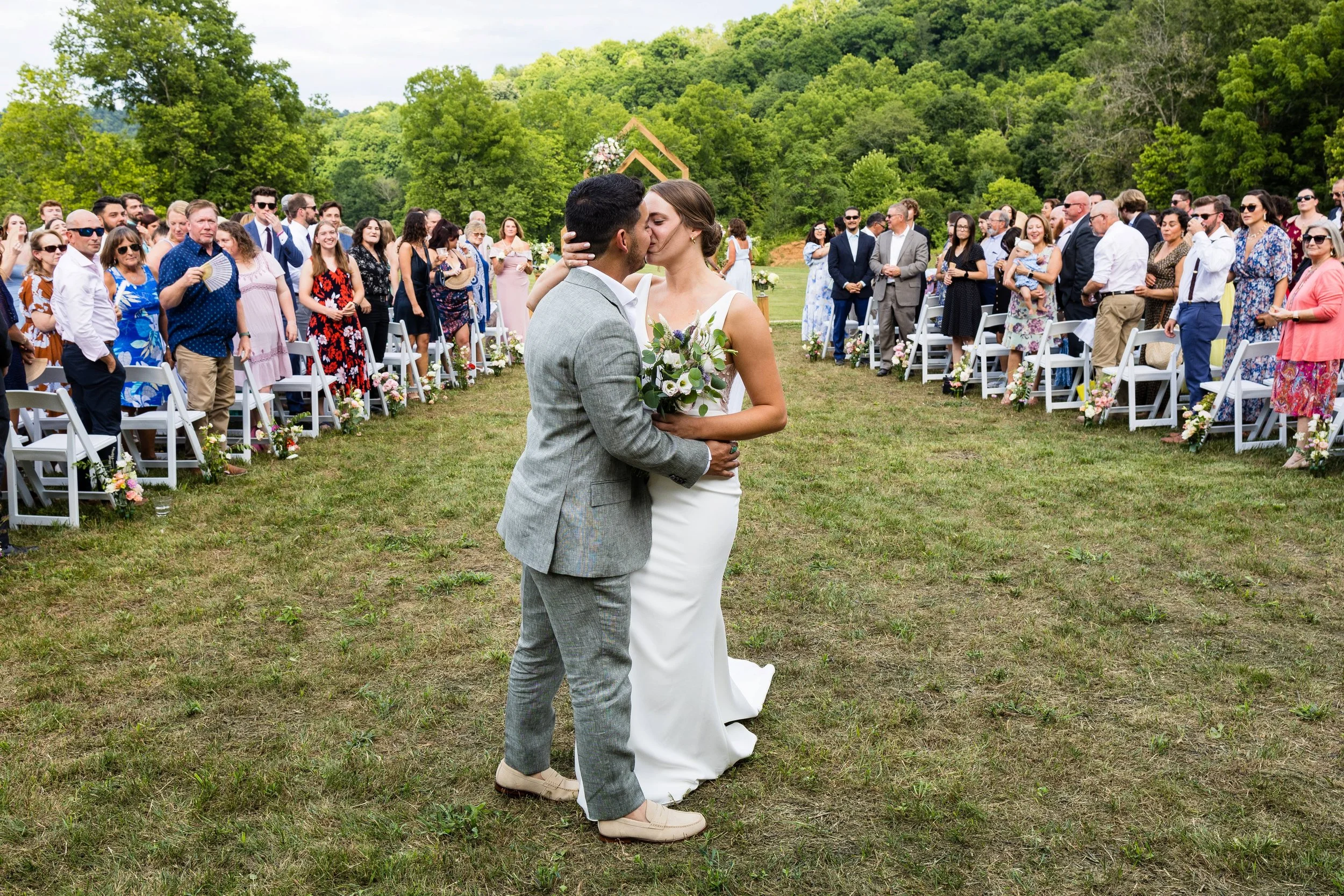 Couple sharing a kiss during their outdoor wedding ceremony, surrounded by guests seated on white chairs decorated with flowers, in a lush green outdoor setting.