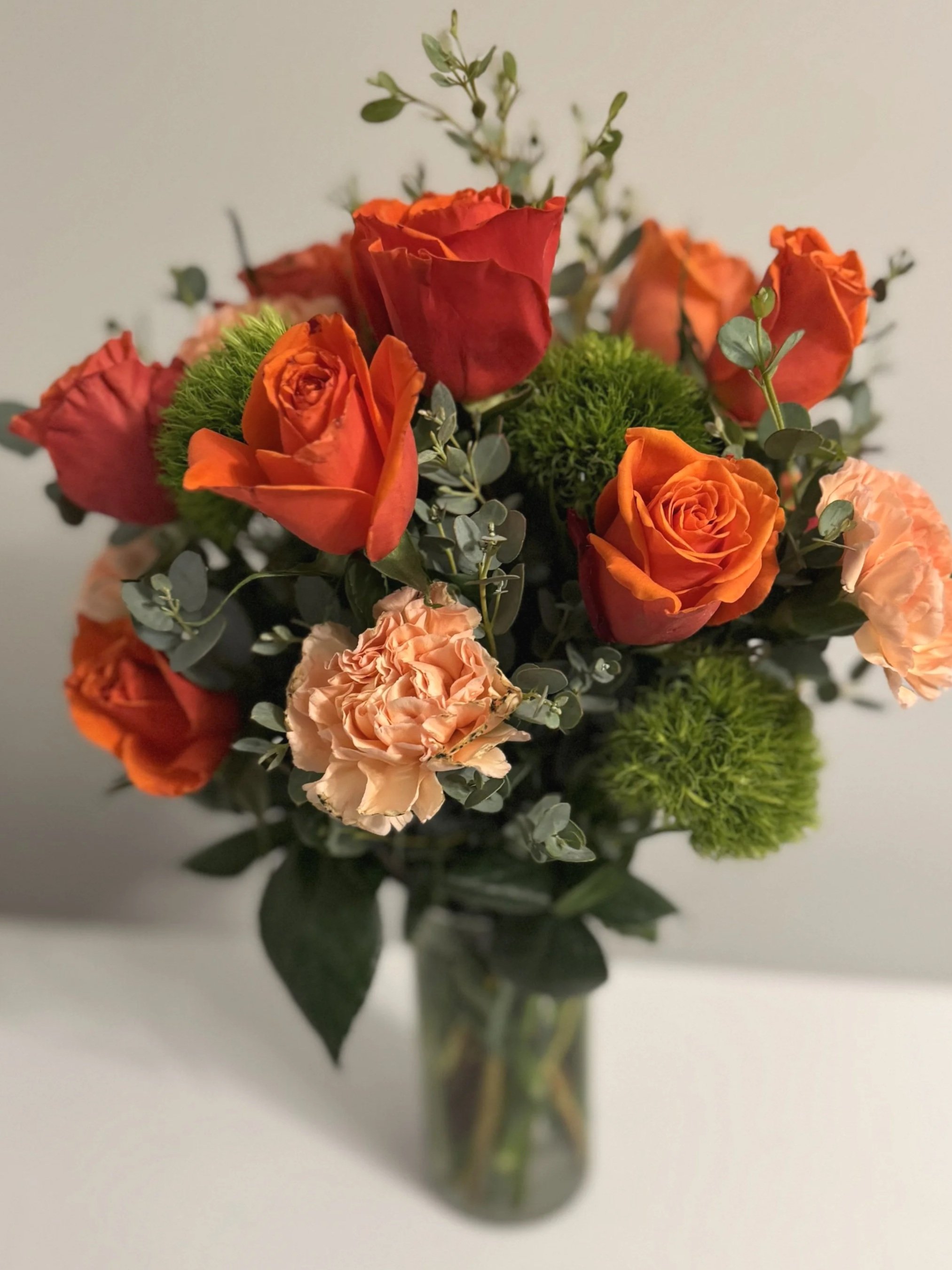 Orange roses and peach carnations with eucalyptus fill a clear cylinder vase on a white table.