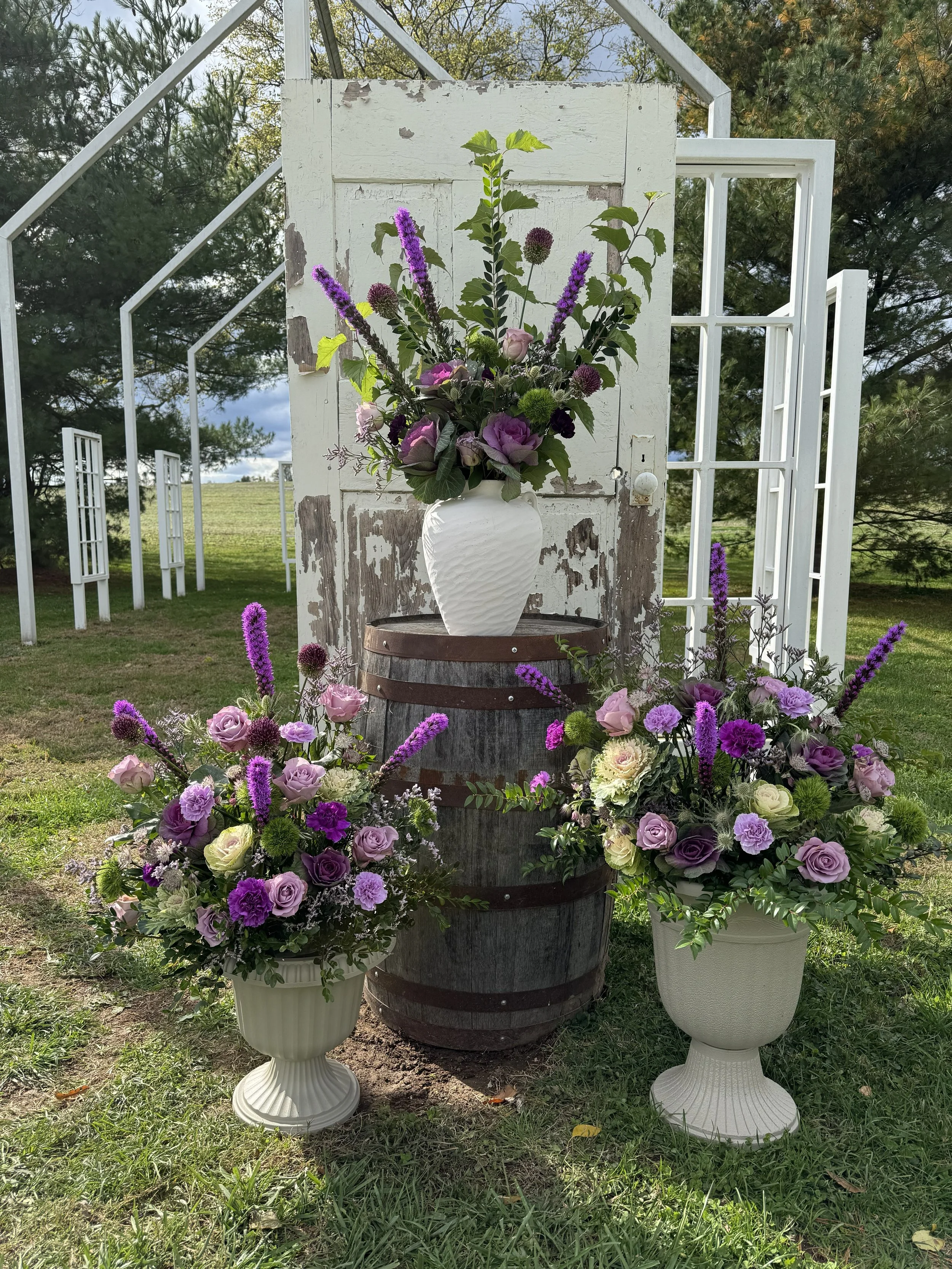 Decorative floral arrangement in white vases and on a wooden barrel, with a rustic white door and outdoor setting in the background.