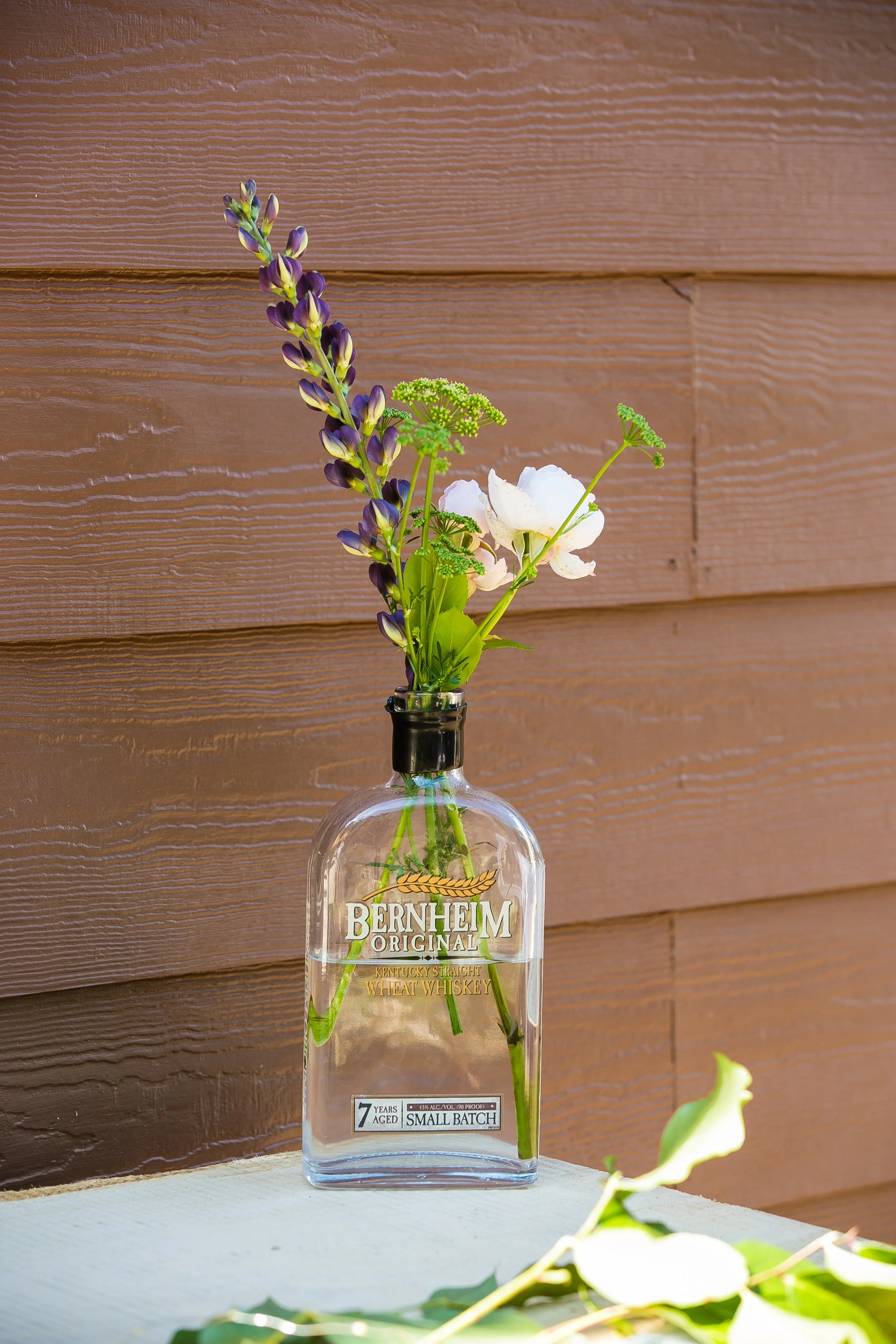 A clear glass bottle labeled Bernheim Original Kentucky Straight Wheat Whiskey used as a vase with purple, white, and green flowers, placed on a white surface against a wooden wall background.