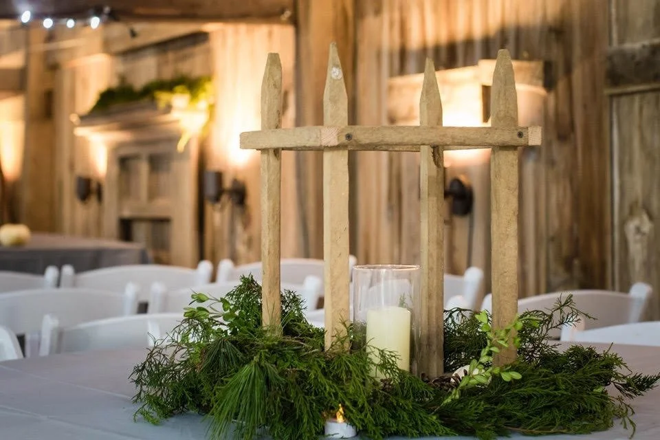 Decorative centerpiece with a wooden fence, a candle in a glass holder, and greenery on a banquet table inside a rustic wooden venue.
