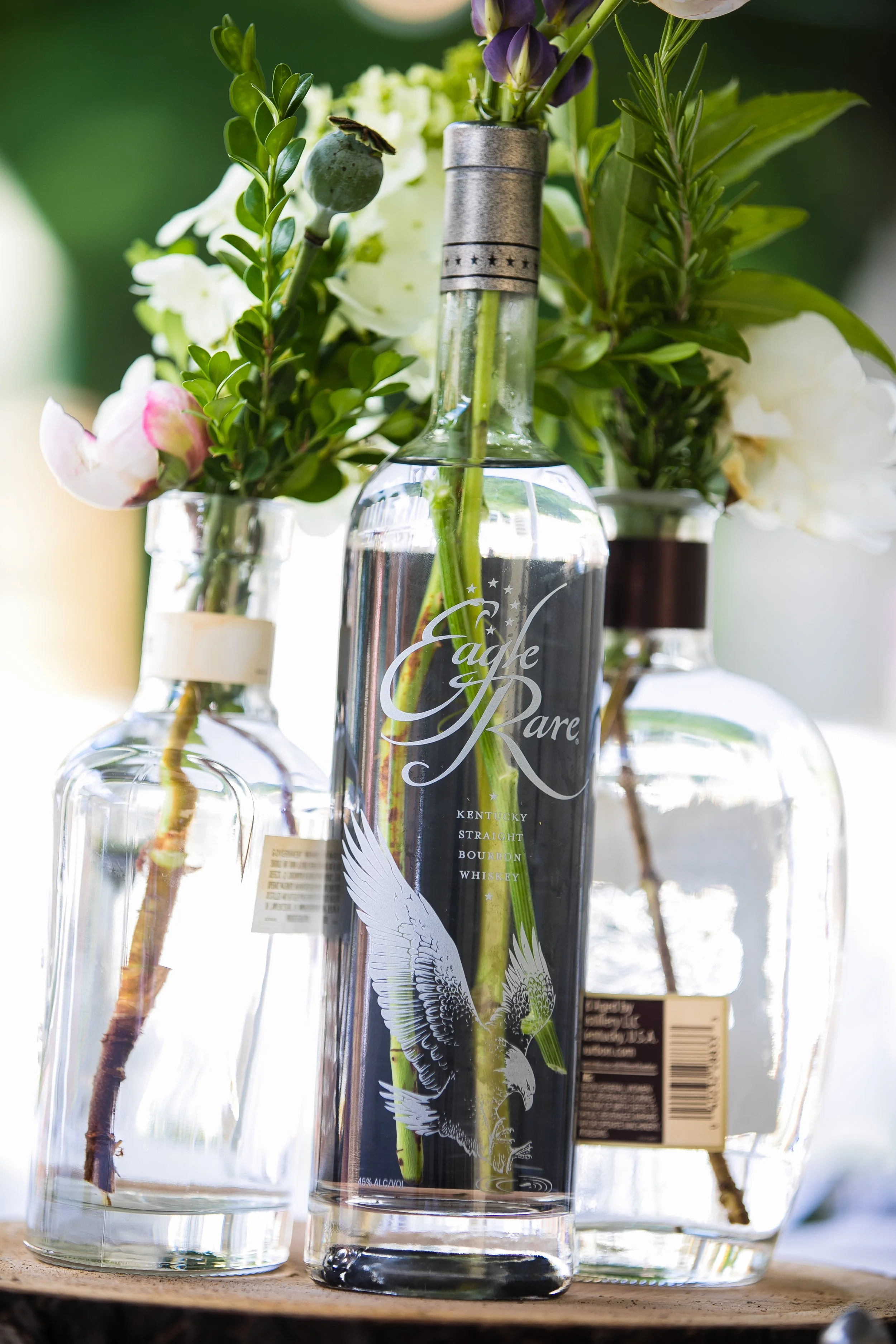 Three glass bottles with flowers arranged inside them, one of which is a vodka bottle with a label featuring a flying eagle.