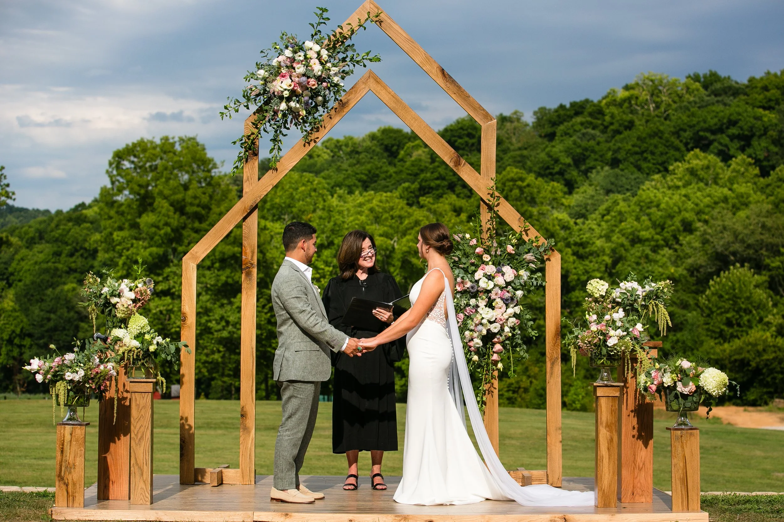 A bride and groom holding hands during their outdoor wedding ceremony, with an officiant smiling in between them, under a wooden arch decorated with flowers, against a background of green trees and cloudy sky.