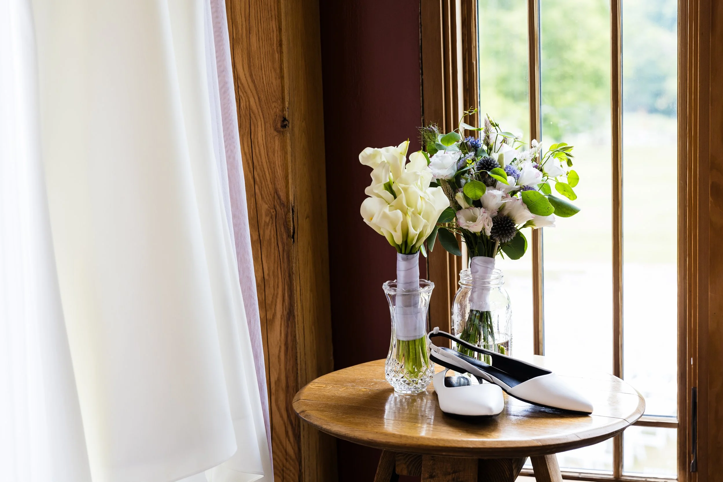 A wooden table by a window with white curtains, holding two glass vases with flowers and a pair of white and black shoes.