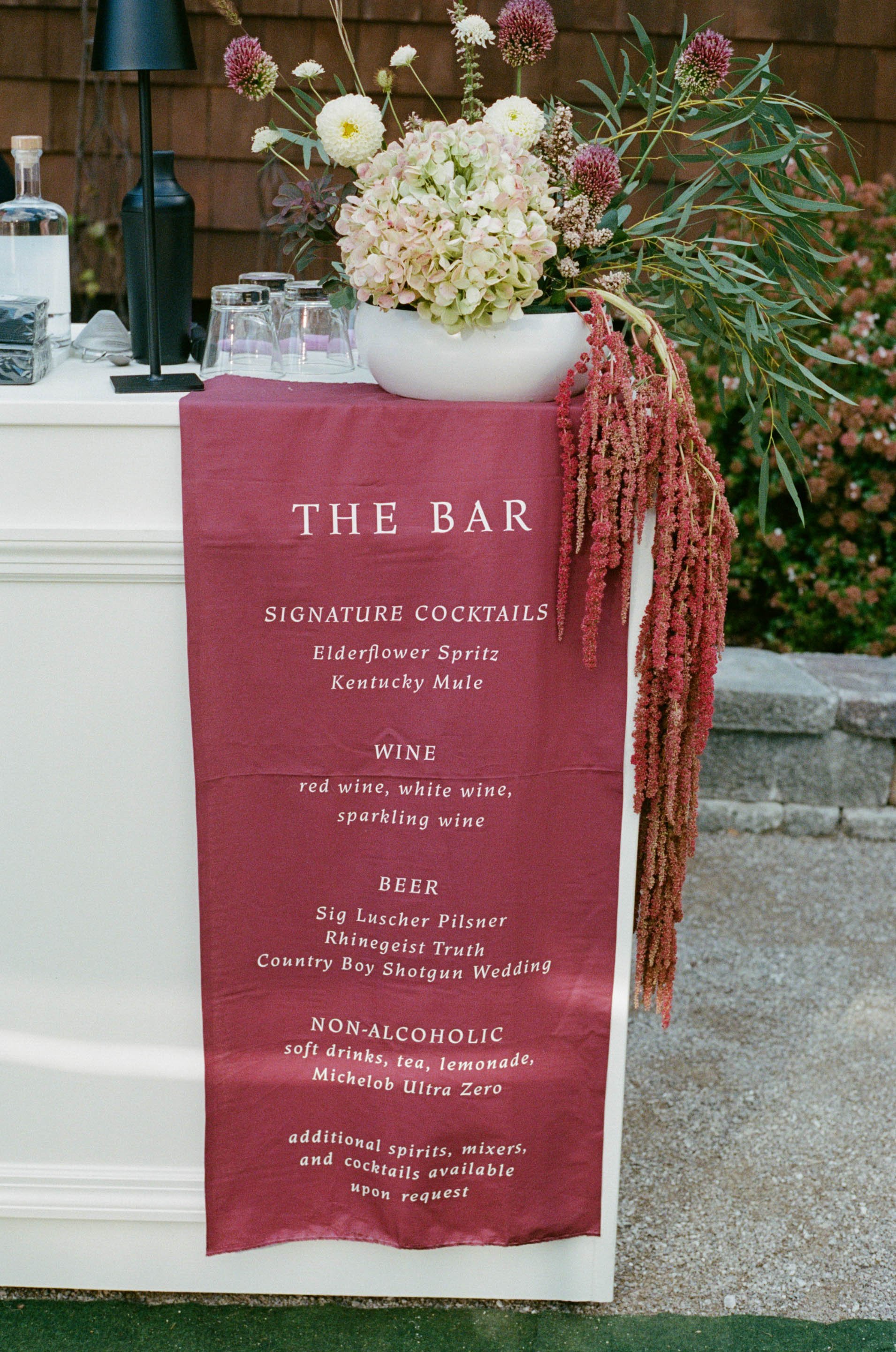 Outdoor bar setup with a wine and cocktail menu, decorated with a large flower arrangement of hydrangeas, alliums, and other flowers, with glasses and drinks on the bar.