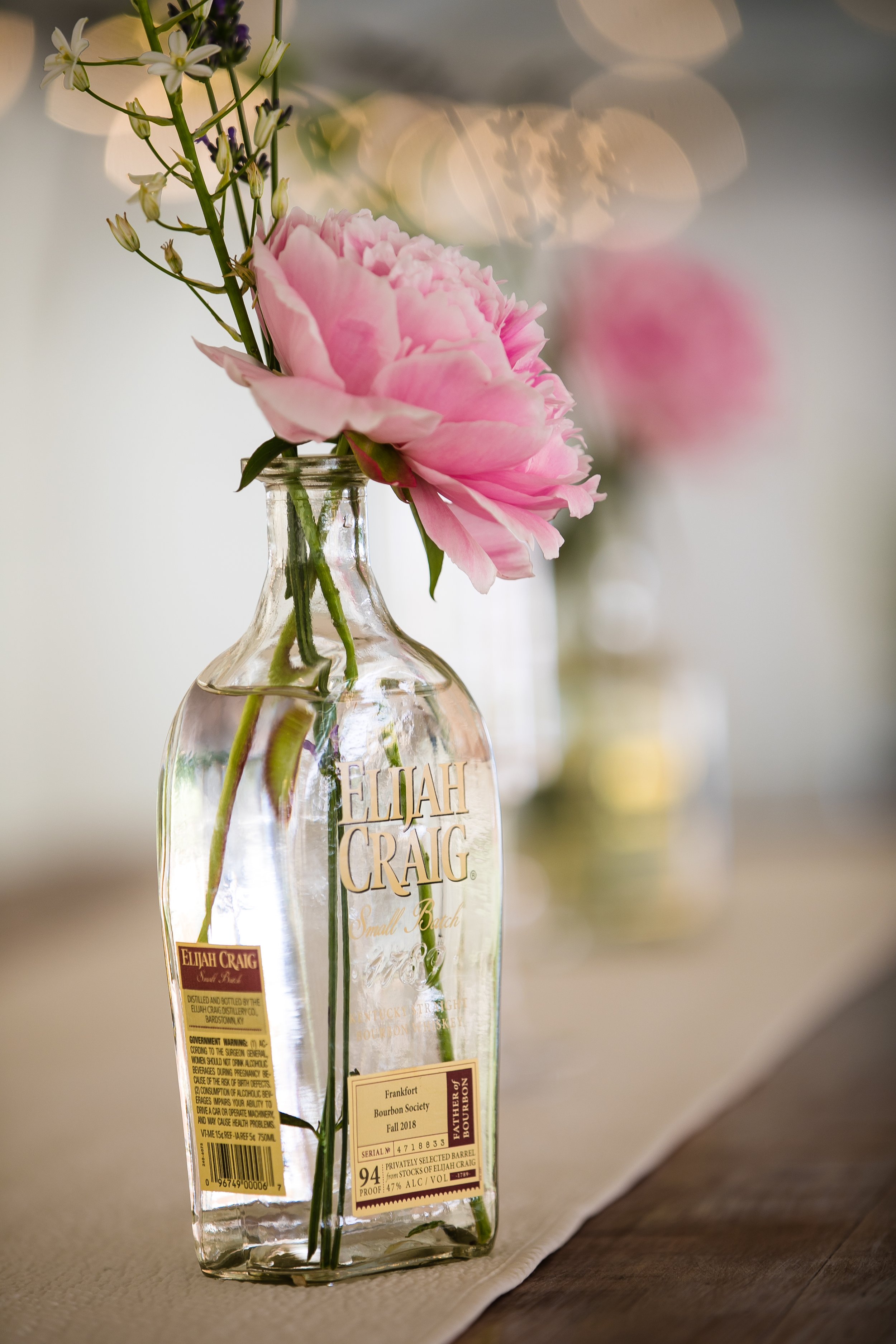 Pink peony and purple flowers in a clear Elijah Craig whiskey bottle used as a vase on a table.