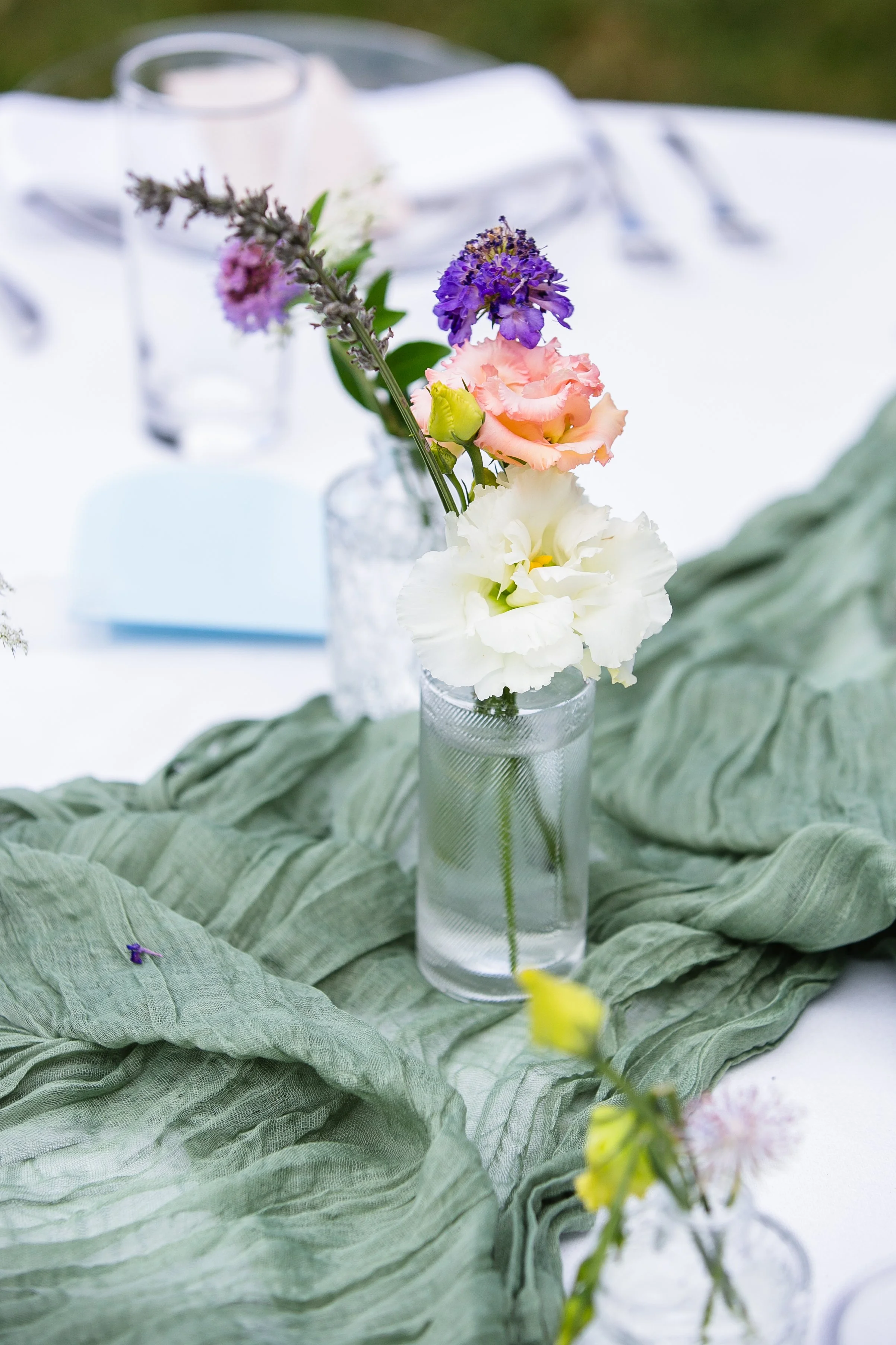 A floral centerpiece with pink, white, purple, and yellow flowers arranged in a clear glass vase on a table with a green fabric runner.