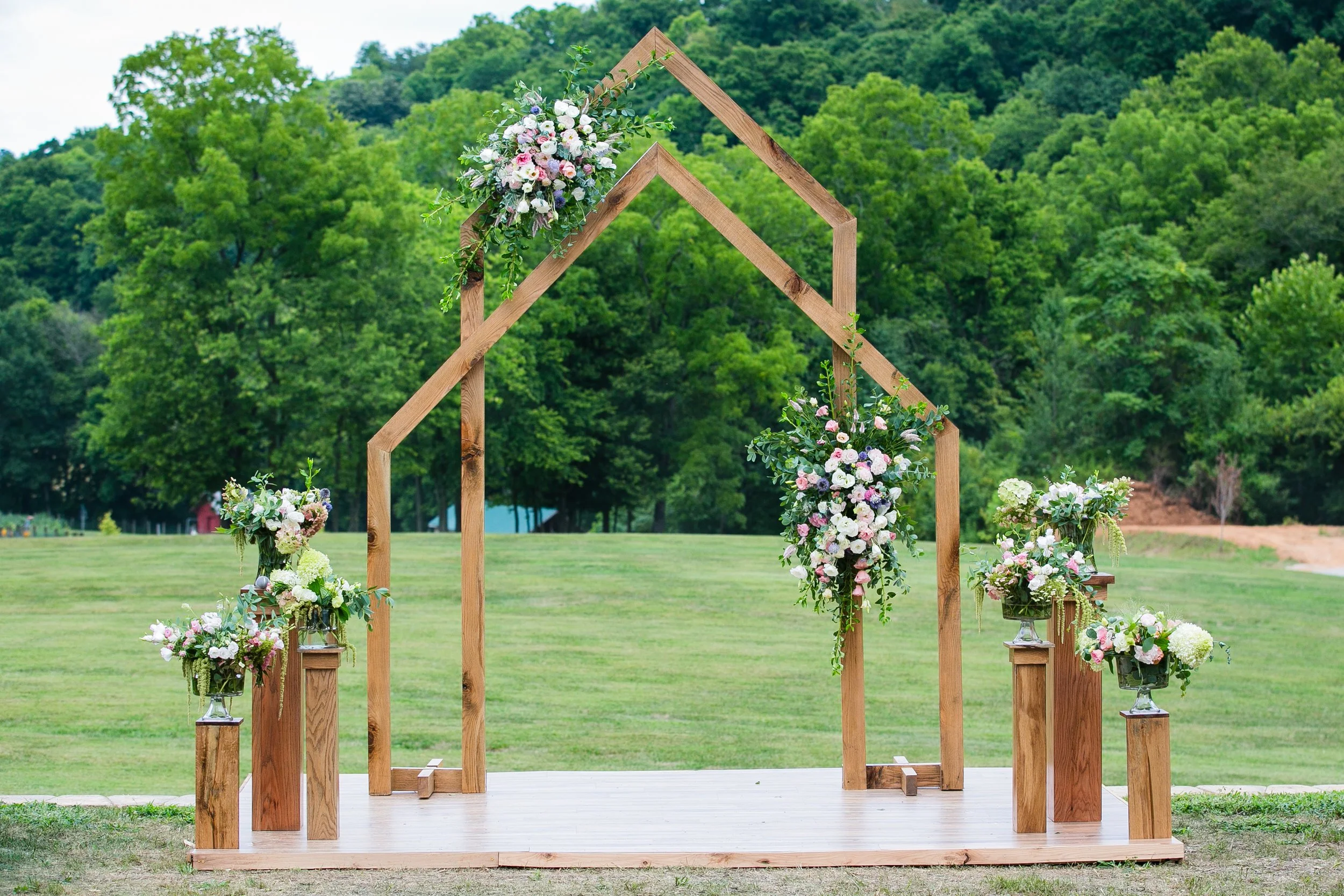 Wooden wedding arch decorated with pastel-colored flowers and greenery, set on a small wooden platform in an outdoor green field with trees in the background.