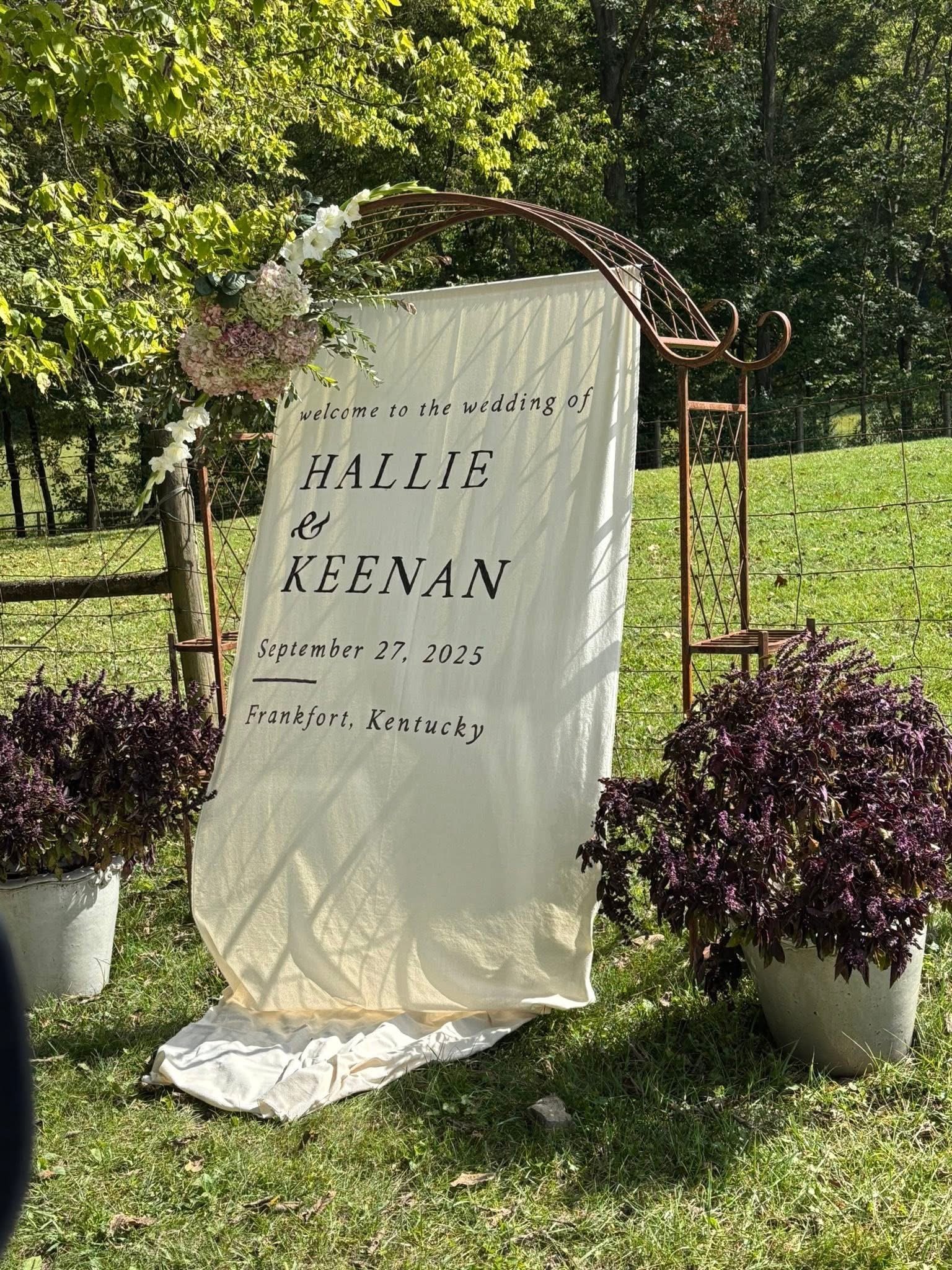 Outdoor wedding welcome sign with floral decorations, announcing the wedding of Hallie and Keenan on September 27, 2025, in Frankfort, Kentucky, surrounded by greenery and a wire fence.