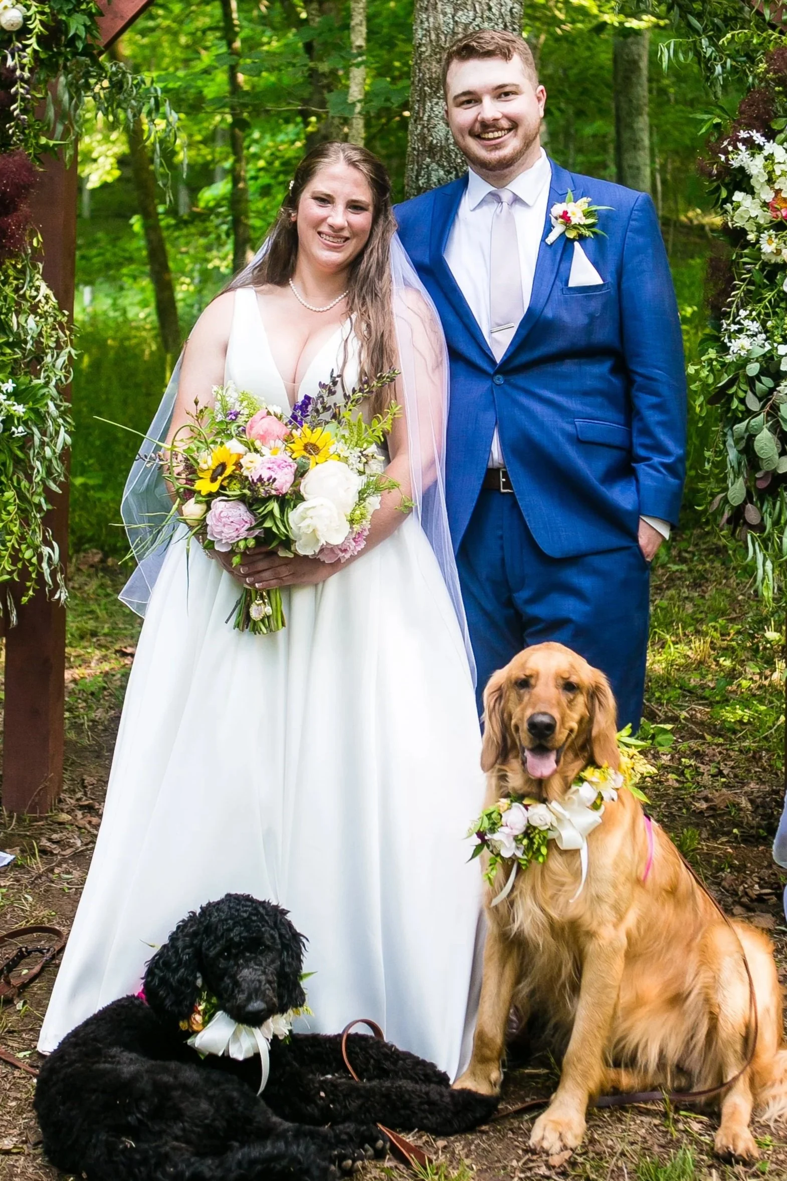 A newlywed couple is standing outdoors in a forested area during their wedding. The bride is wearing a white gown and holding a bouquet of flowers, while the groom is dressed in a blue suit with a boutonniere. They are smiling and standing alongside two dogs wearing floral collars, one black and one golden.
