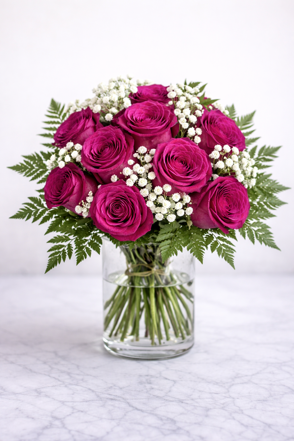 Bouquet of vibrant pink roses with white baby's breath and green fern leaves in a clear glass vase on a marble surface.