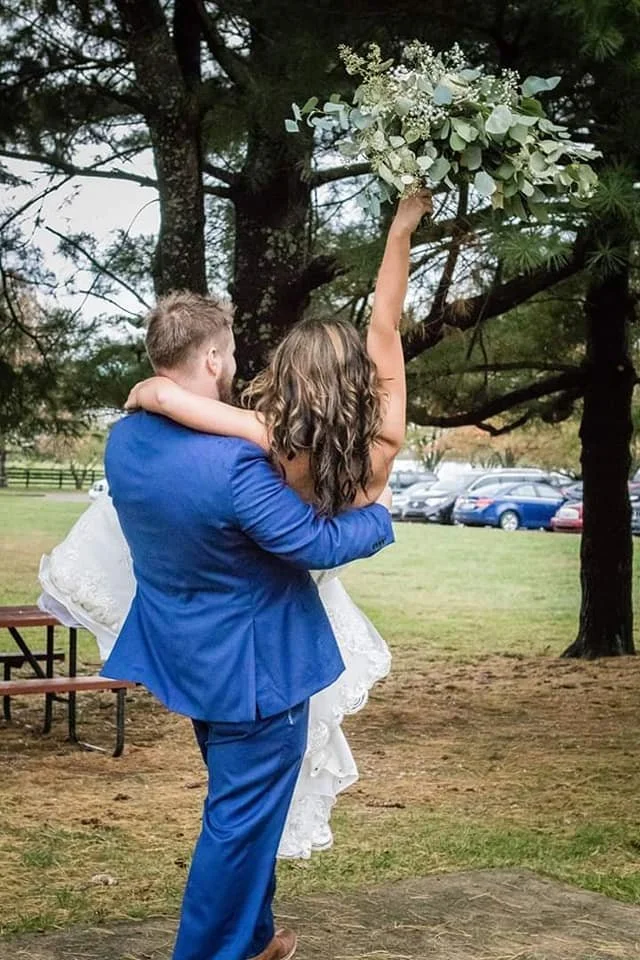 A man in a blue suit is holding a woman in a white dress, lifting her into the air outdoors near trees and parked cars. The woman is reaching up with one arm holding a large bouquet of white flowers and greenery.
