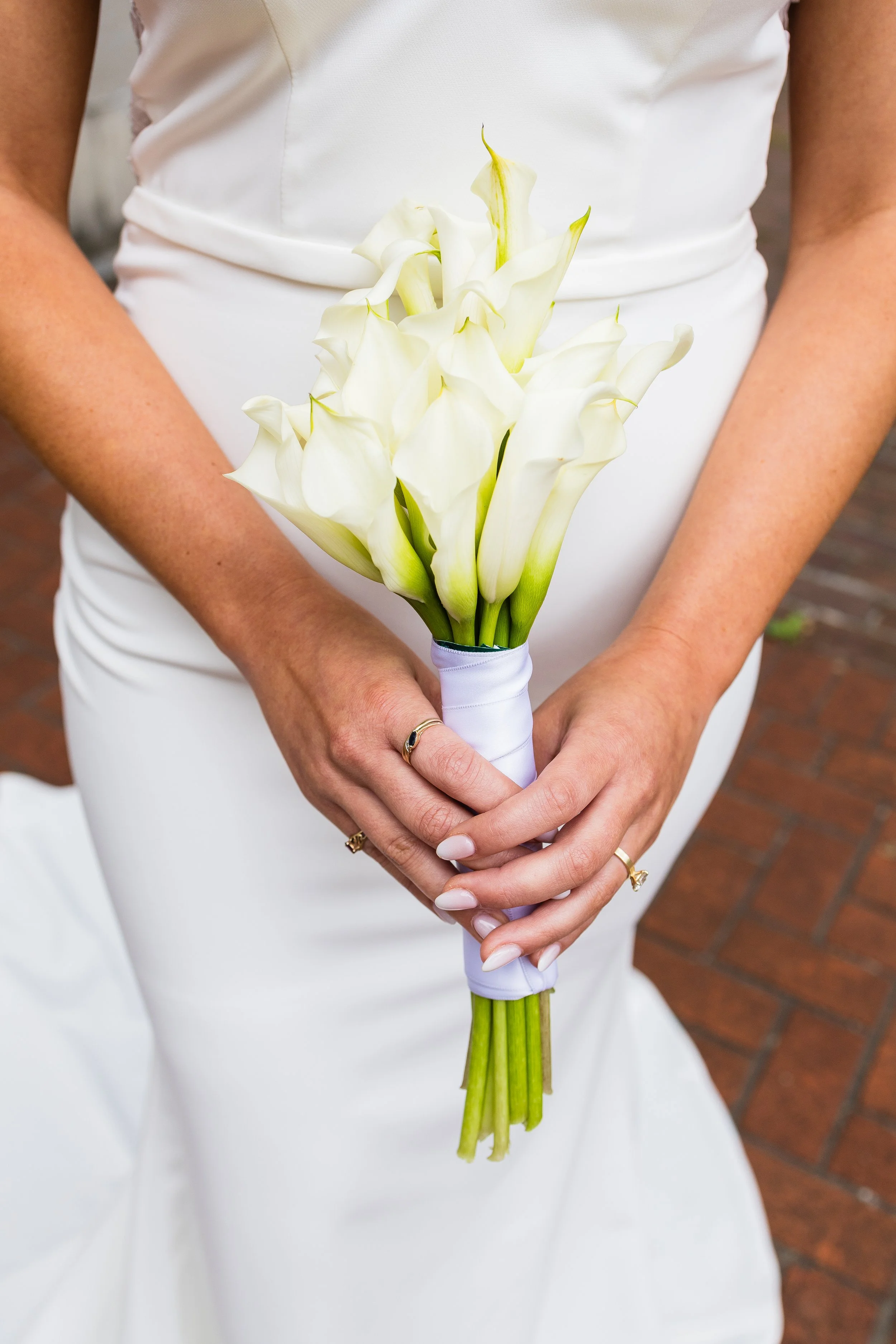 Close-up of a woman in a white dress holding a bouquet of white calla lilies, standing on brick pavement.