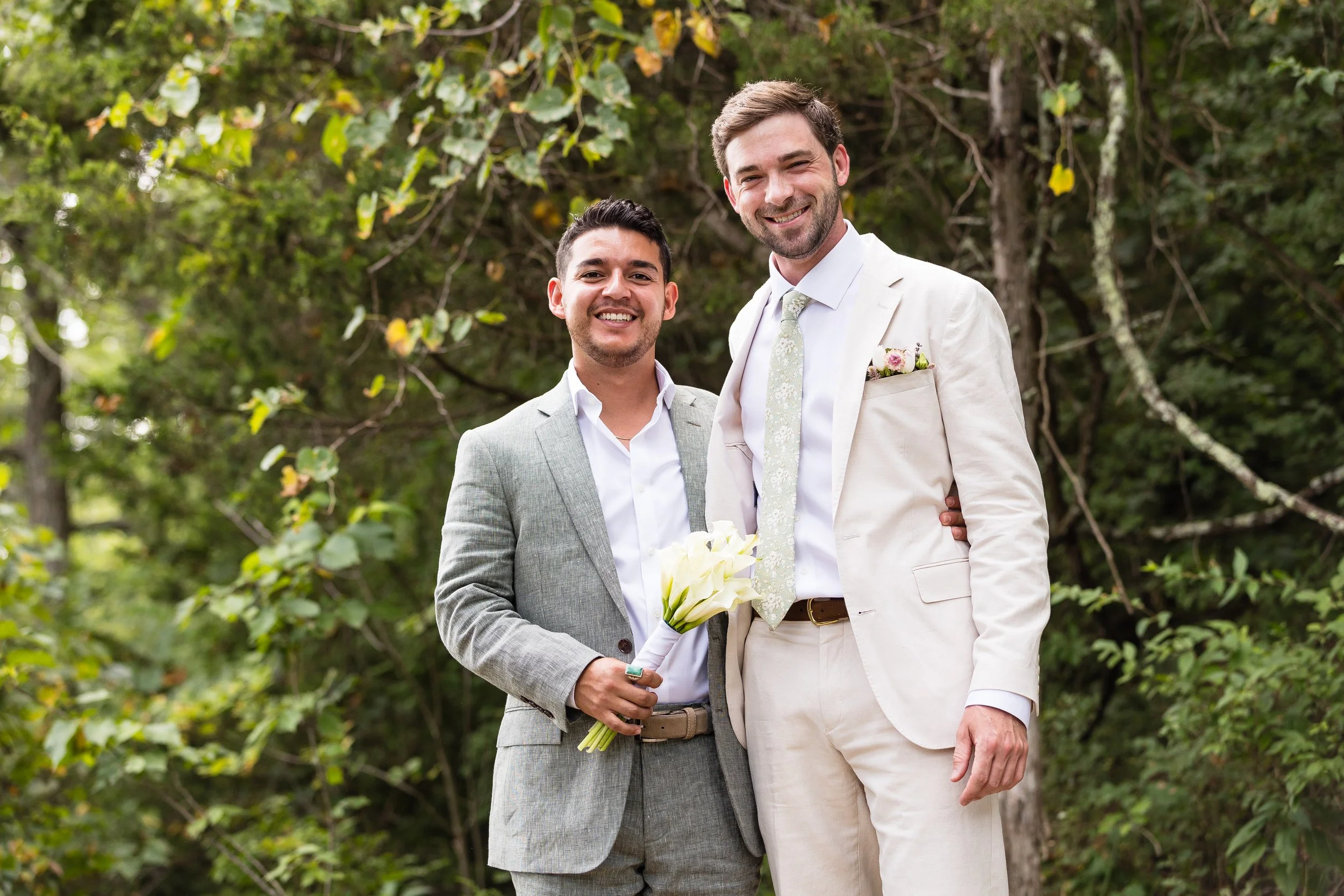 Two men in suits smiling, one holding a bouquet of white flowers, standing outdoors with trees in the background.