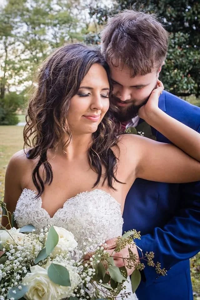 A bride in a white lace wedding dress holding a bouquet of white flowers, standing outdoors with a groom in a blue suit leaning in close and embracing her.