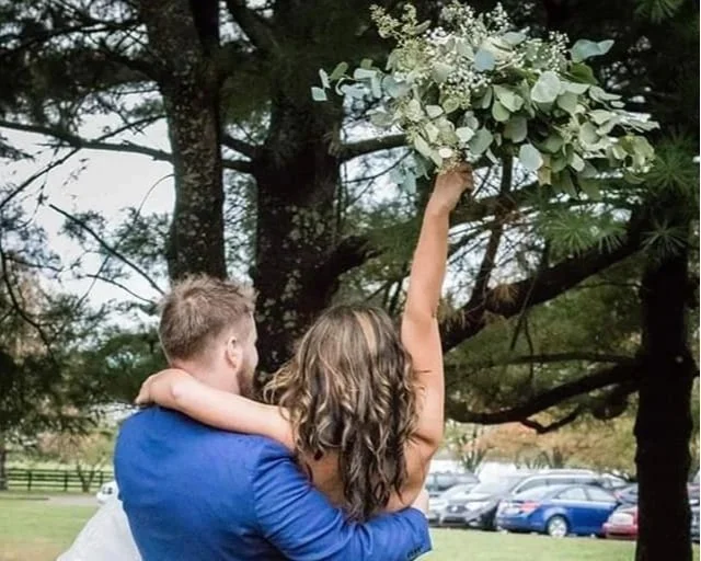 A woman is holding a bouquet of white flowers above her head while sitting on a man's shoulder outdoors, surrounded by trees and parked cars.