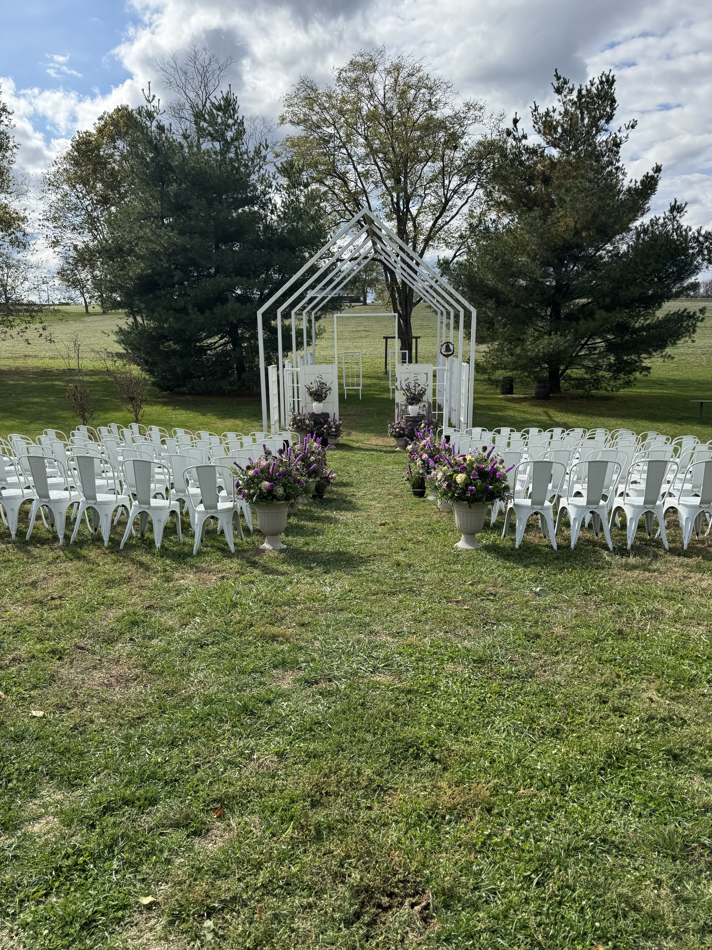 Outdoor wedding ceremony setup with a white wooden arch, arranged chairs, and floral decorations on a grassy field with trees in the background under a partly cloudy sky.