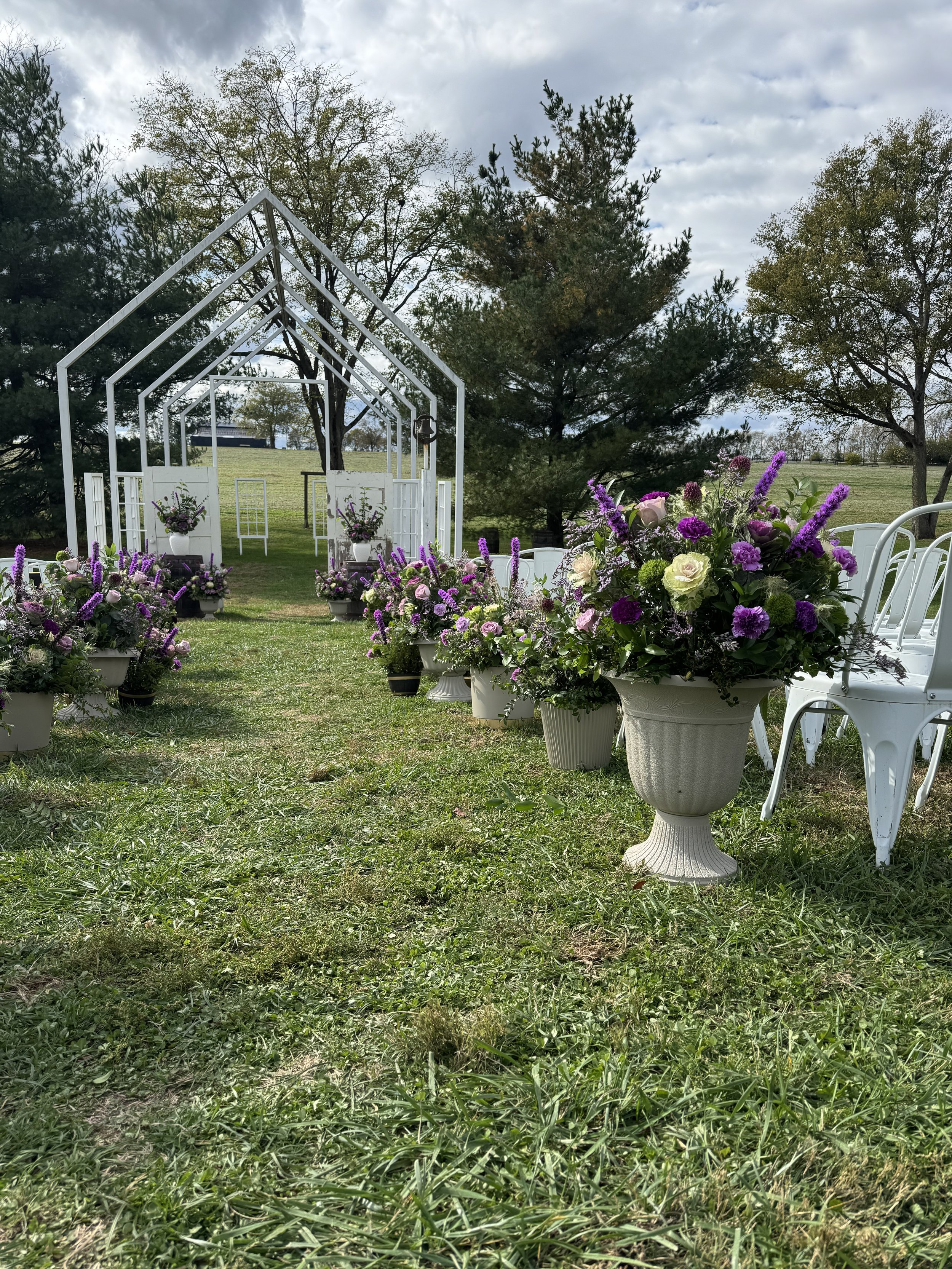 White outdoor wedding arch decorated with purple and white flowers and surrounded by large white planters filled with similar flowers on grass, with trees and a cloudy sky in the background.
