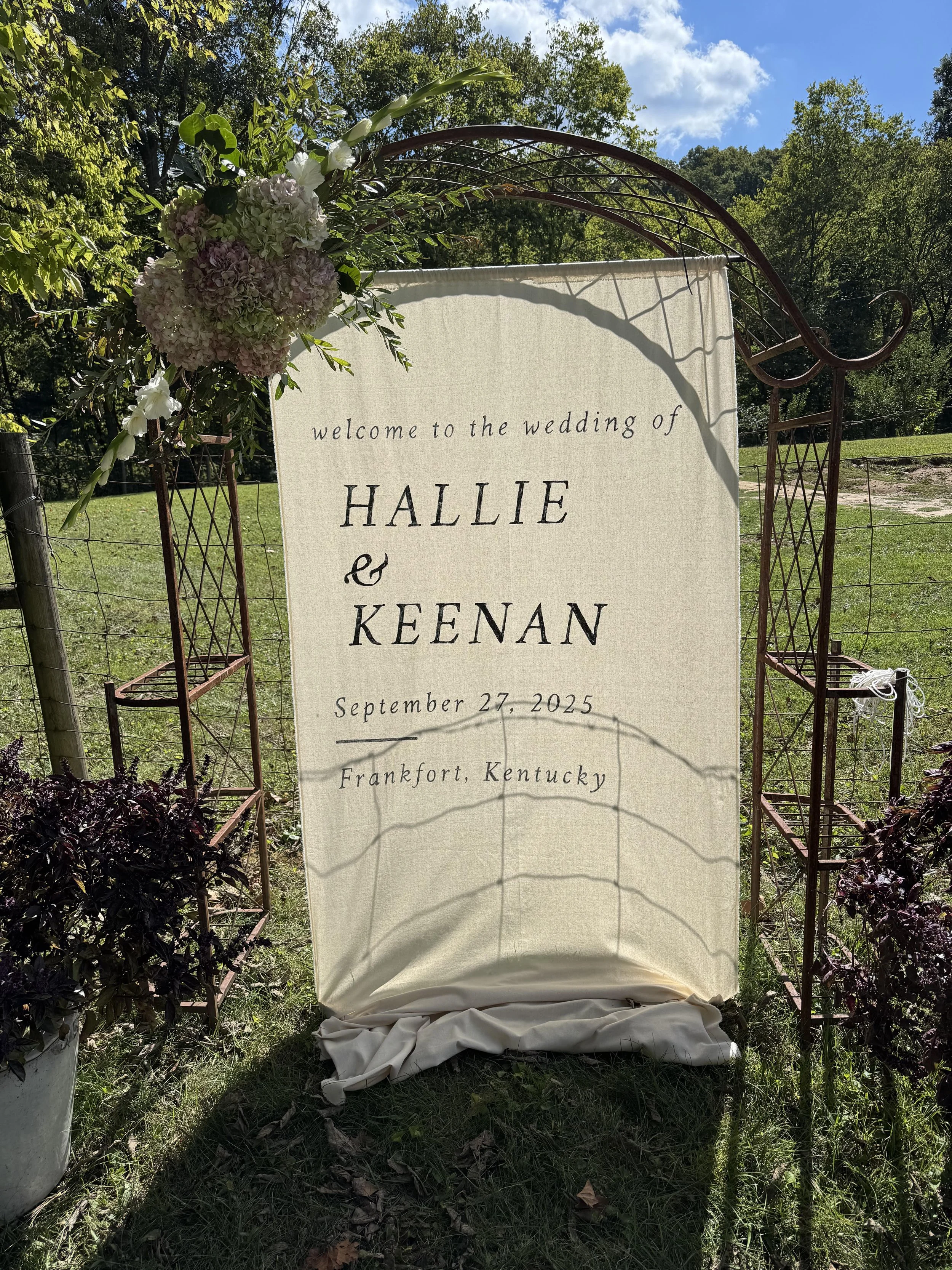 Wedding welcome sign with floral decorations reading, 'welcome to the wedding of HALIE & KEENAN, September 27, 2025, Frankfort, Kentucky' outdoors with trees and a blue sky in the background.