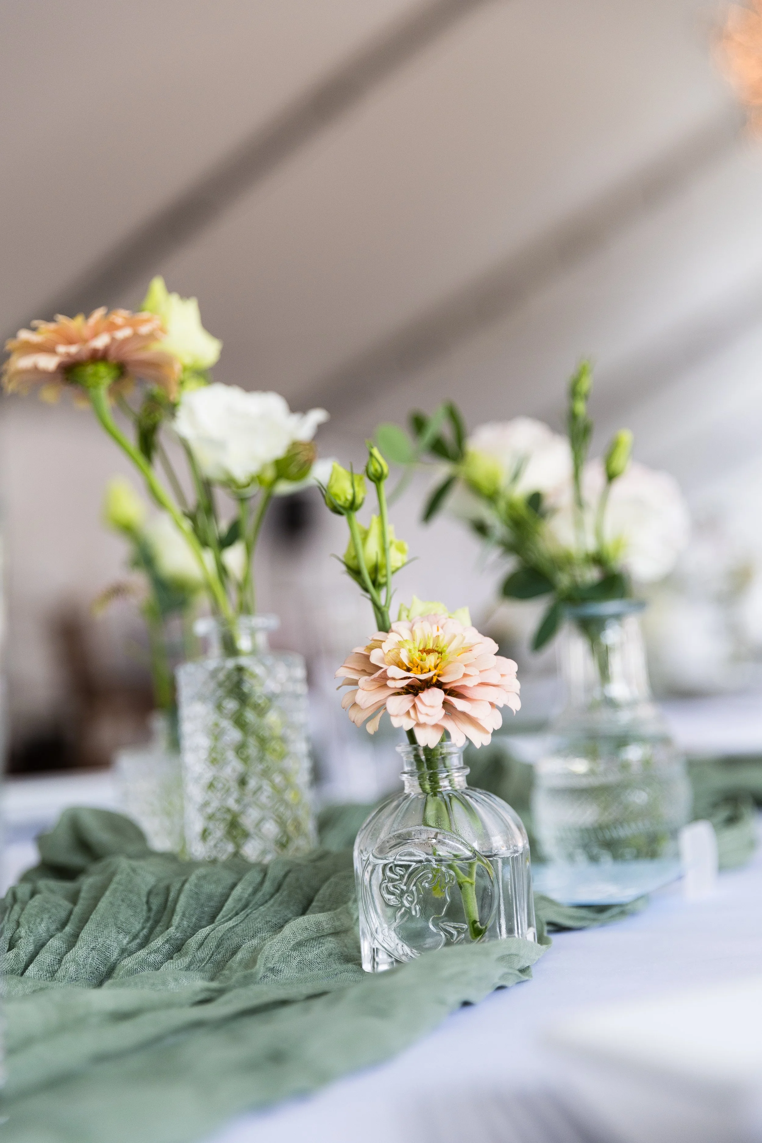 Decorative table centerpiece with pastel-colored flowers in vintage glass bottles on a green fabric runner.