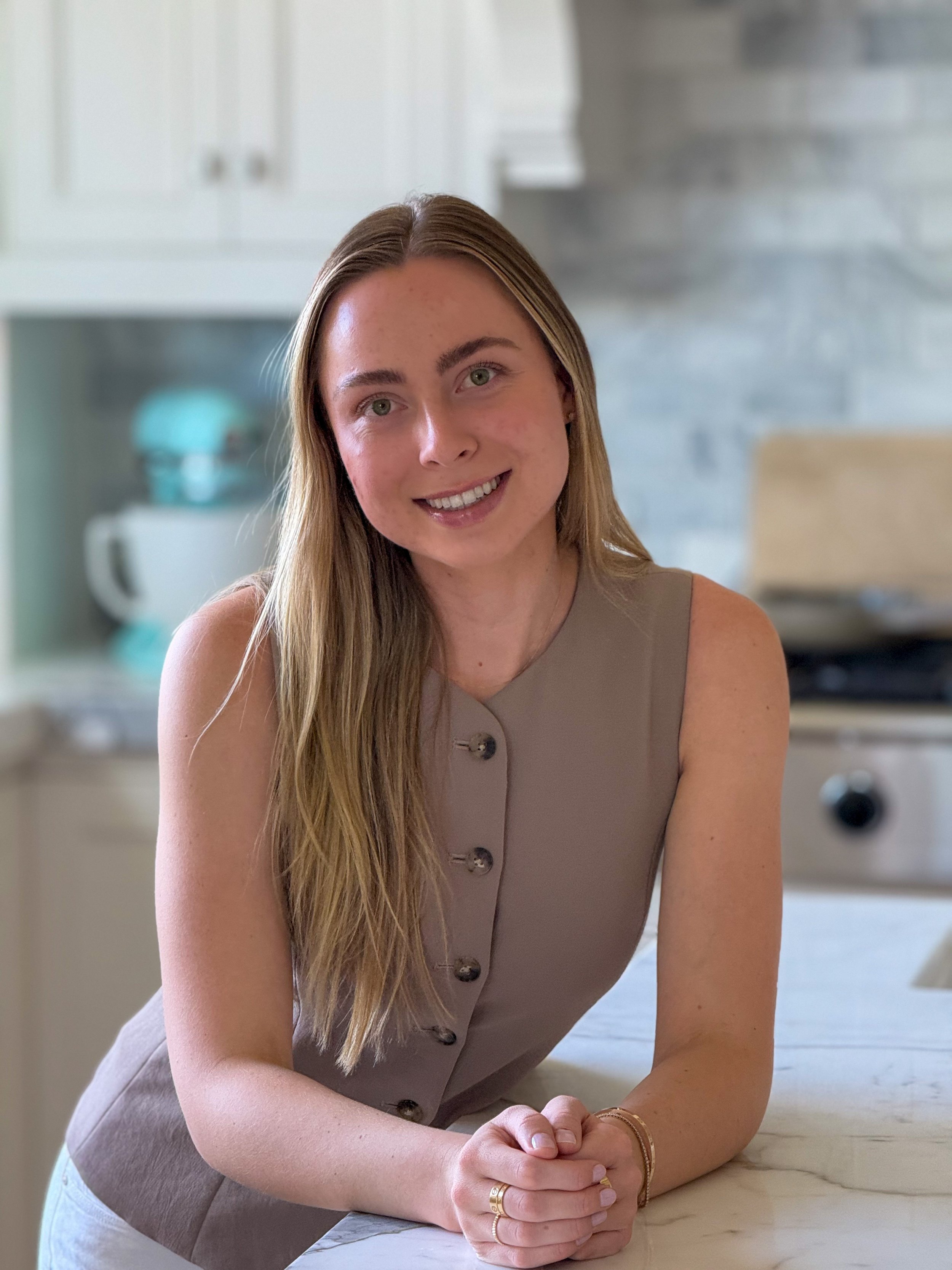 A woman with long blonde hair and green eyes, wearing a sleeveless beige top, smiles and poses with her hands folded on a kitchen counter.