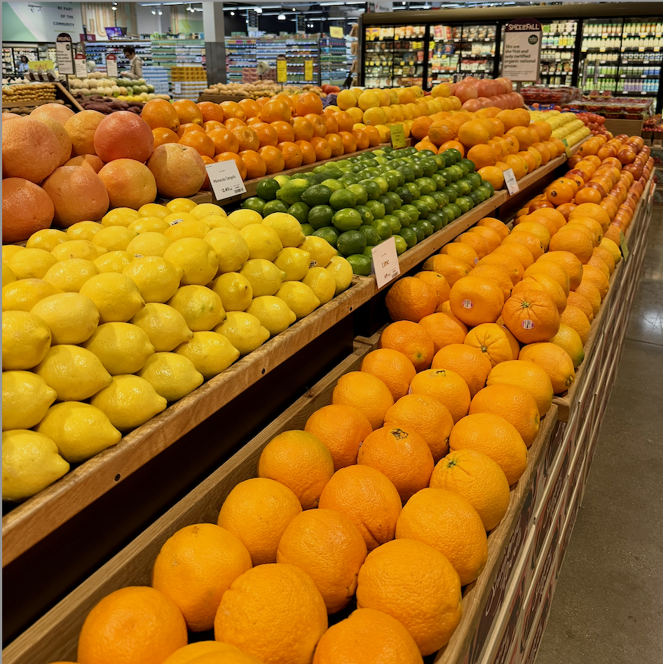Display of fresh citrus fruits including lemons, oranges, grapefruits, apples, and limes arranged on wooden shelves in a grocery store produce section.