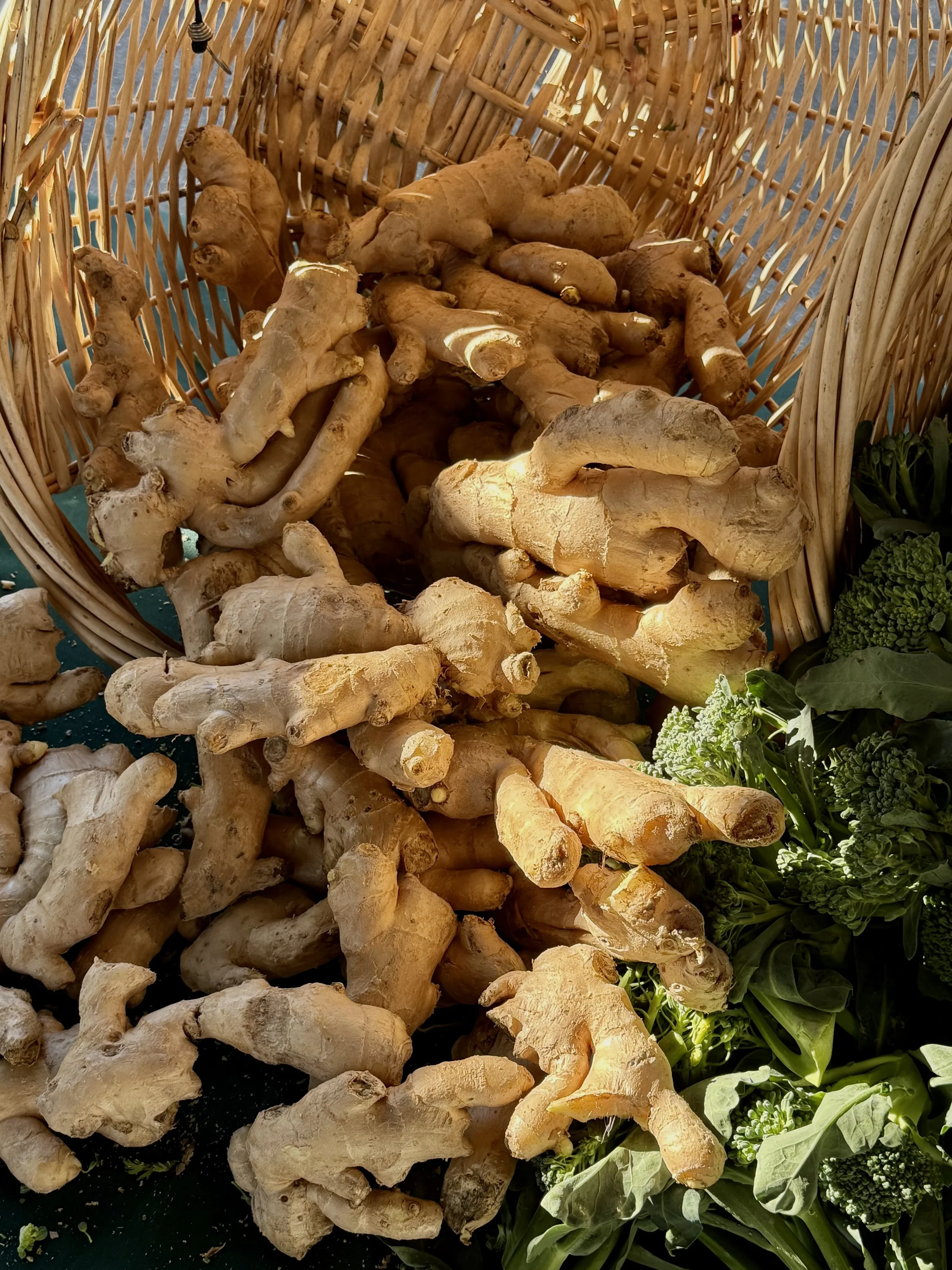 Fresh ginger roots in a basket at a market, with green vegetables nearby.