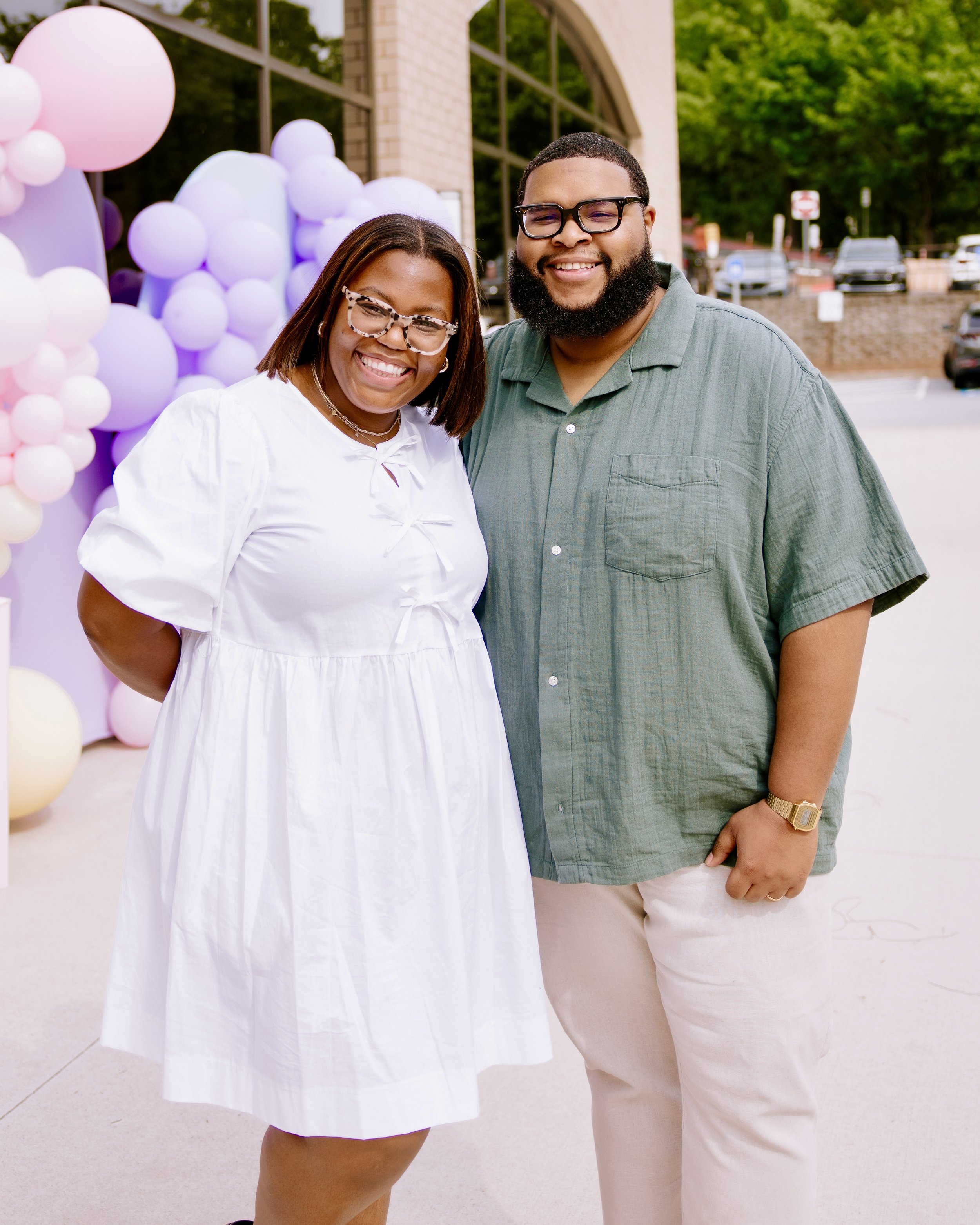 A smiling woman in a white dress with sunglasses and a man with glasses, a beard, and a green shirt, standing outdoors near a building with balloons in the background.
