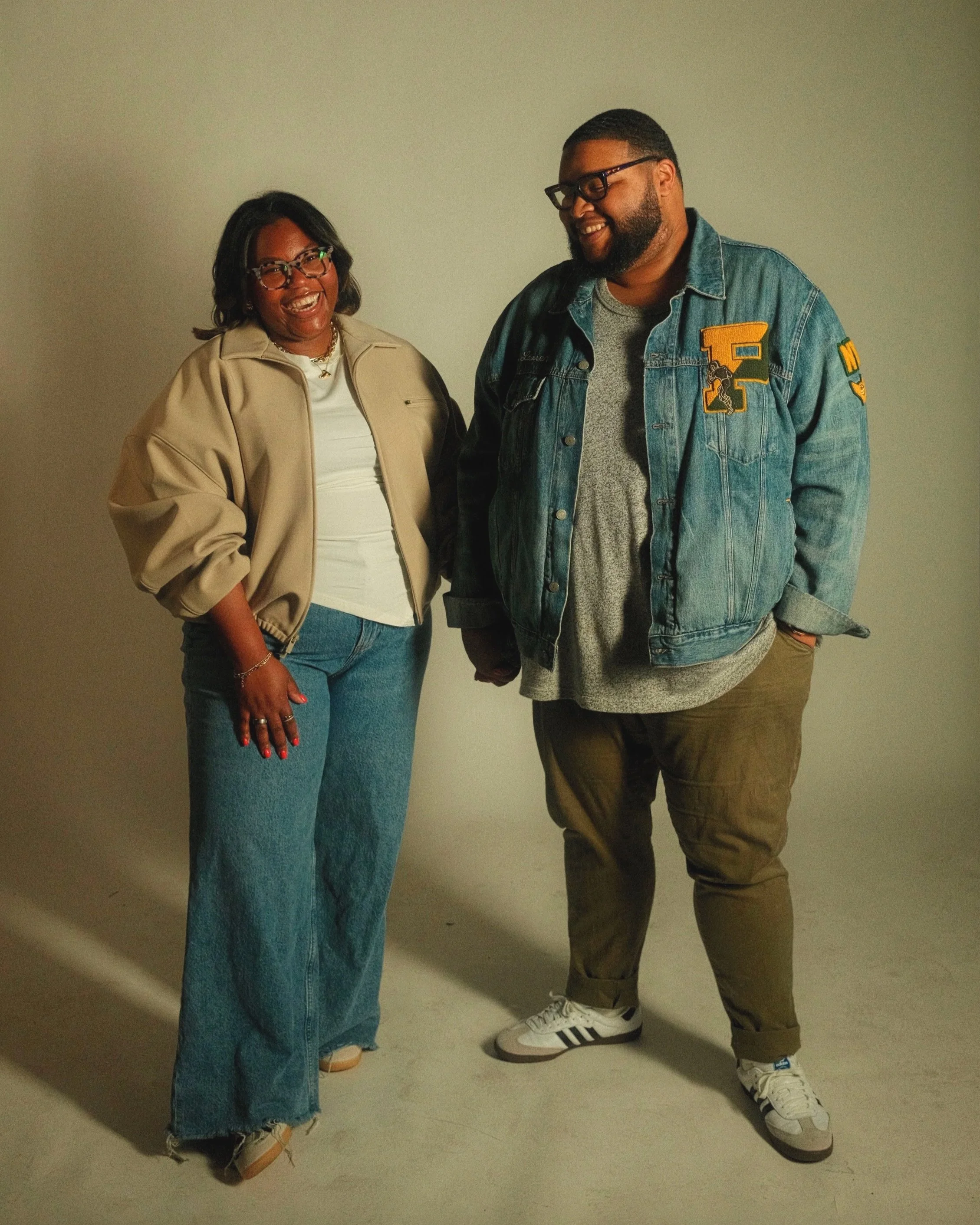 A woman and a man standing together, smiling and holding hands, in a photo studio with a plain background.