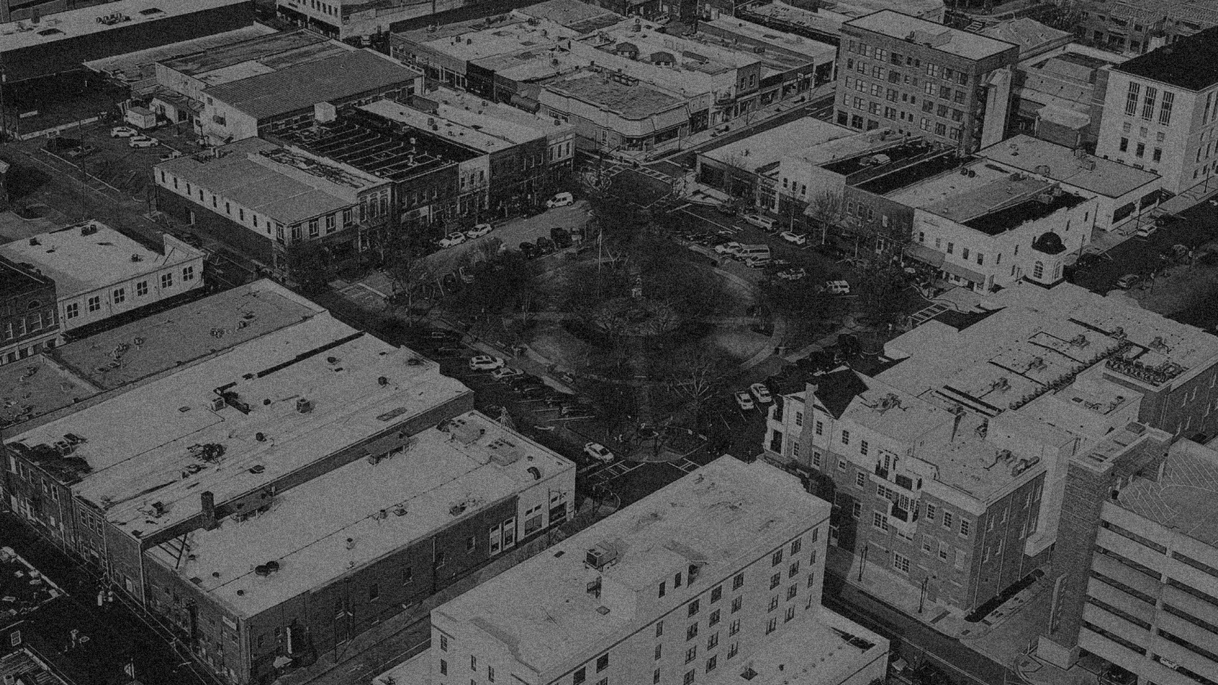 Black and white aerial view of a city block with various buildings, a small park with trees, and parking lots filled with cars.