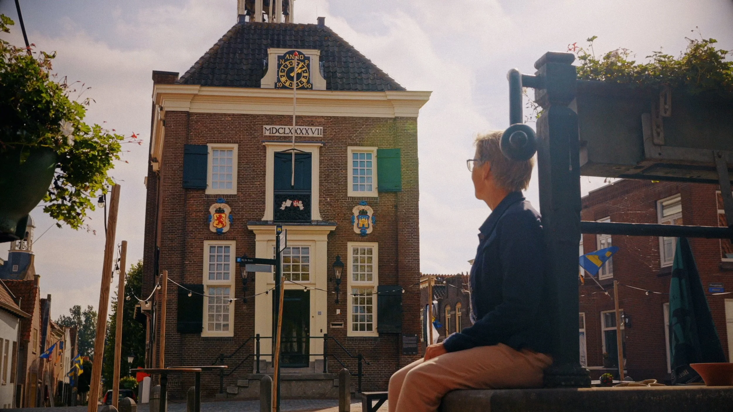 A person with glasses and gray hair sitting on a bench in front of a historic brick building with a clock on the facade and decorative shields, in a European town square during daylight.