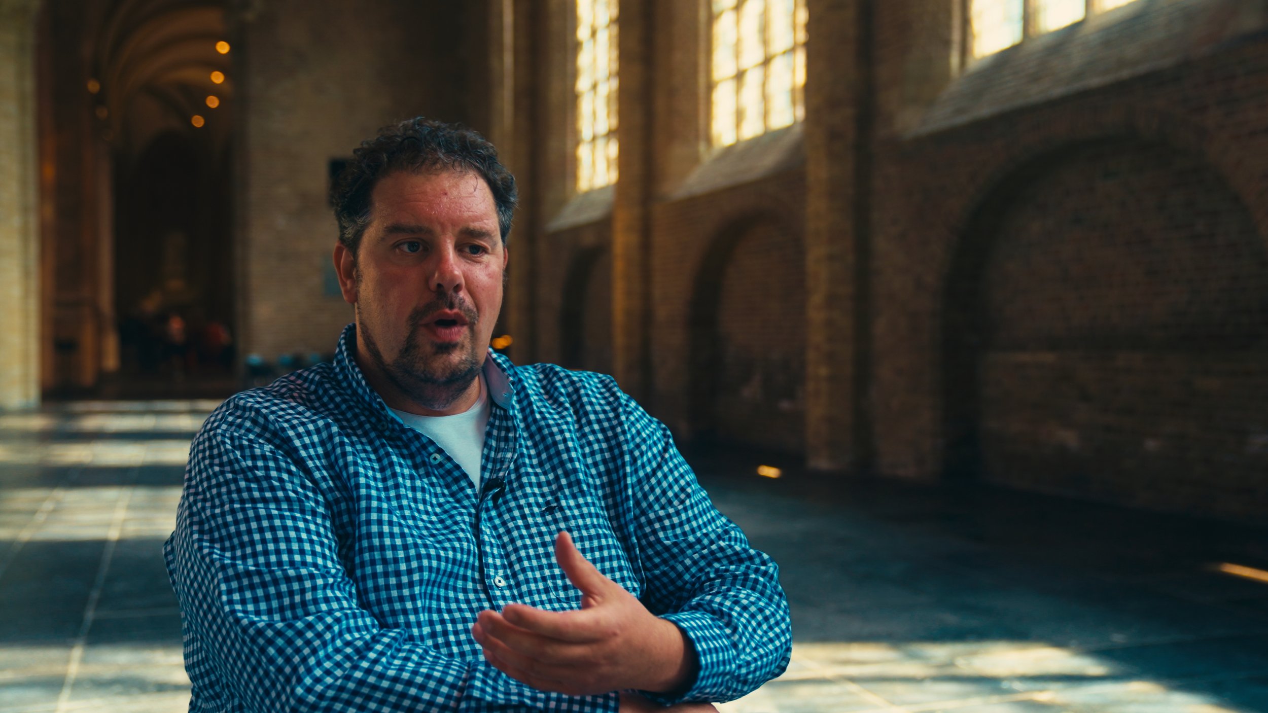 Man with curly hair and beard wearing a blue checkered shirt speaking in a large, historic, brick hall with high arched windows and sunlight streaming through.