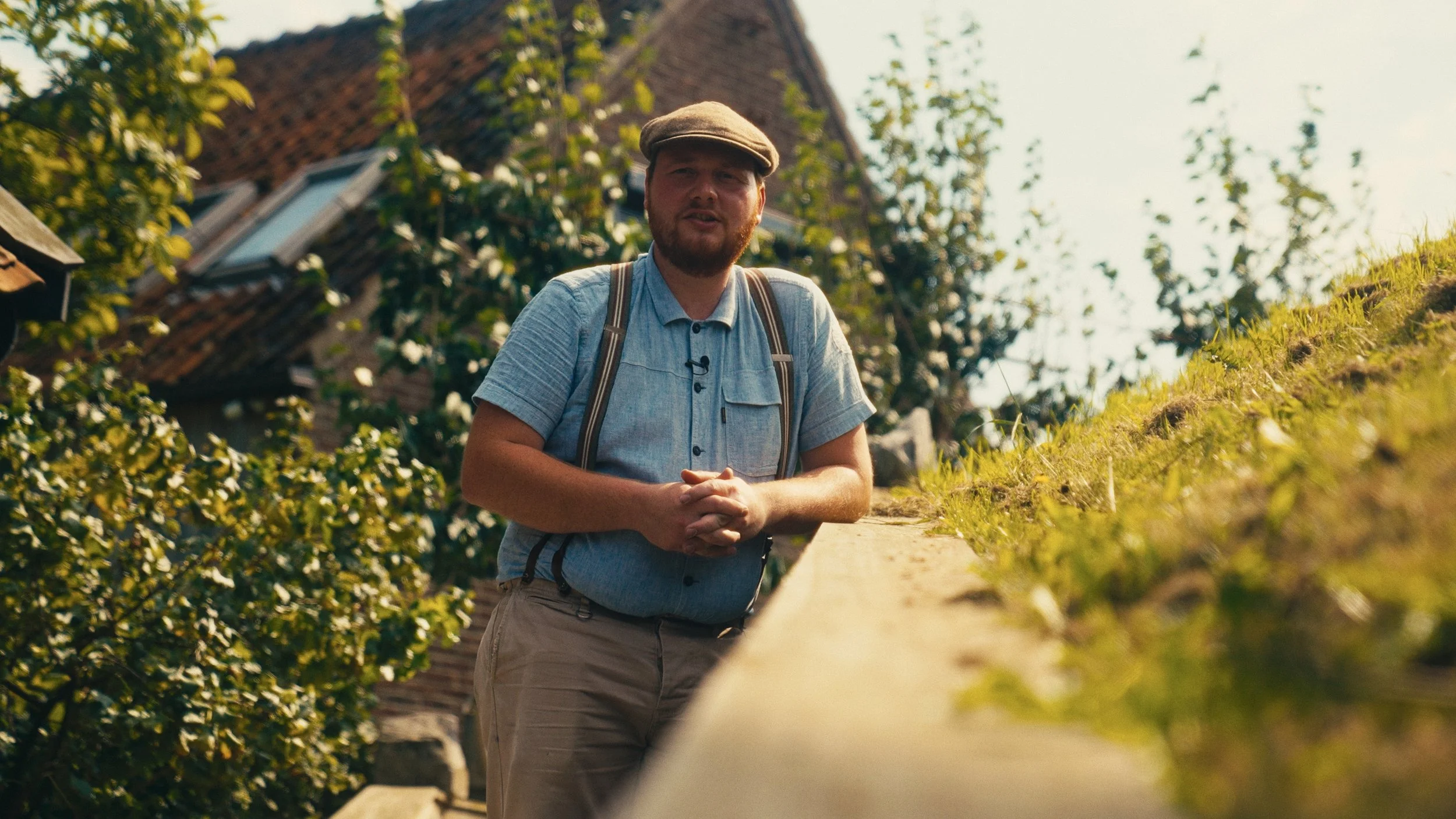 A man with a beard and mustache wearing a cap, light blue short-sleeve shirt, suspenders, and khaki pants, standing outdoors near a stone wall with a house and trees in the background.