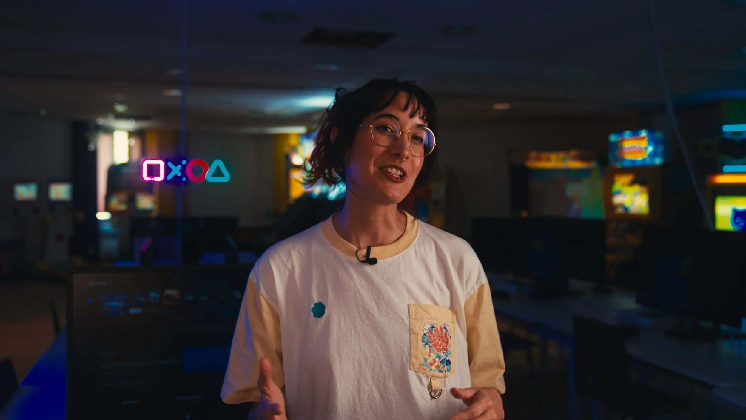 A woman in a well-lit arcade or gaming room, wearing glasses and a cream-colored shirt with a floral patch pocket, smiling and speaking, surrounded by neon gaming and colorful digital art displays.