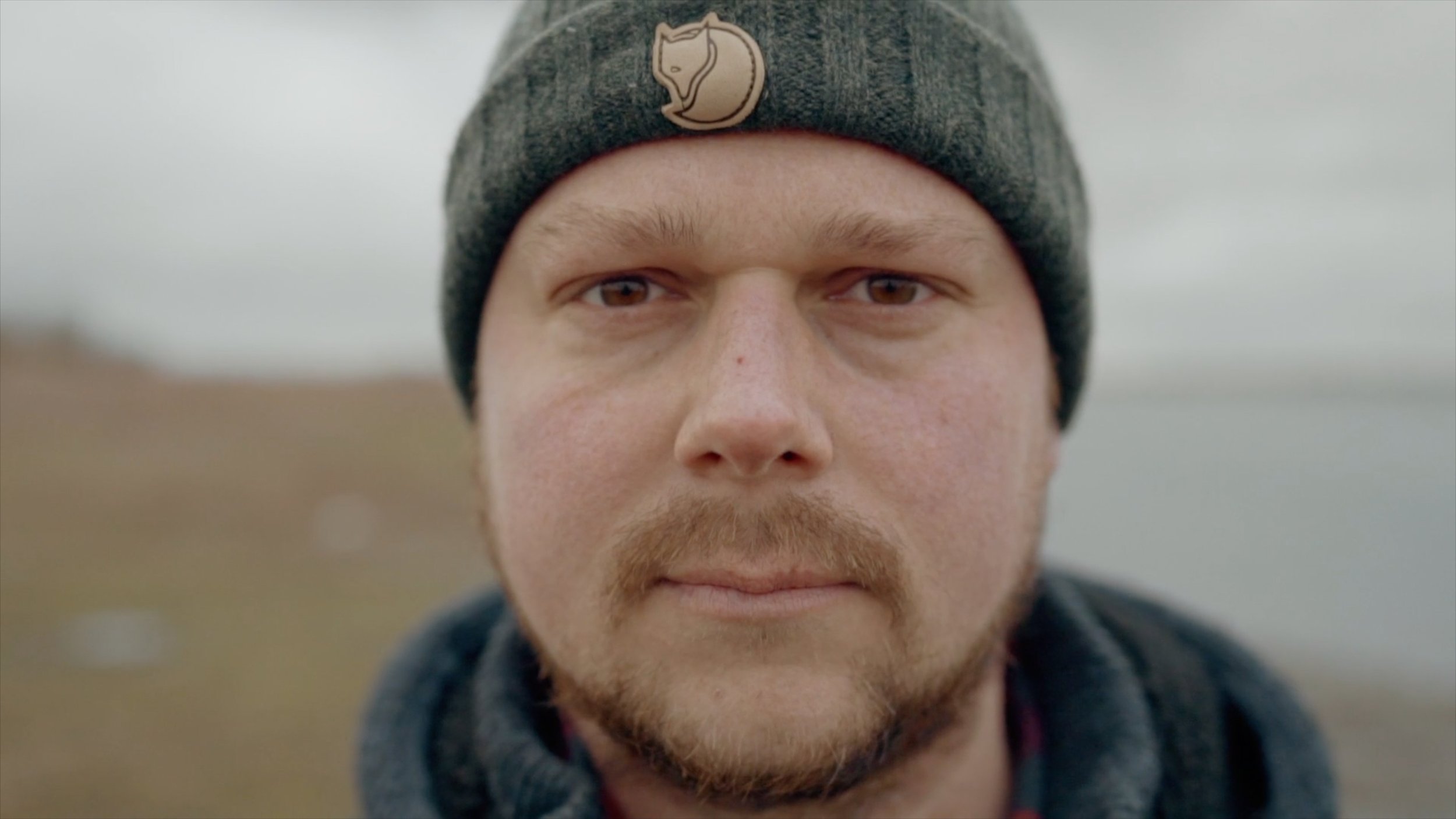 Close-up of a man with light skin, brown eyes, reddish-brown beard, wearing a gray beanie with a small logo and a dark jacket, outdoors in a foggy or overcast environment.