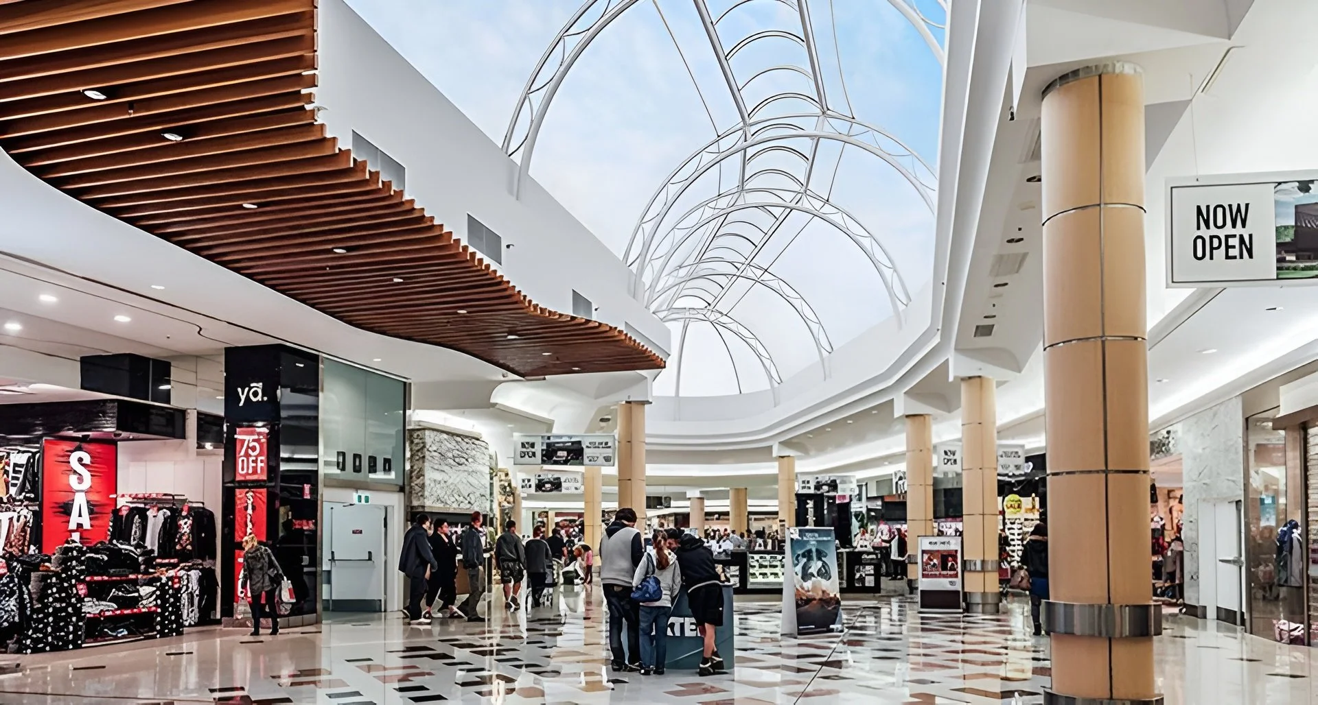 Inside a modern shopping mall with shoppers walking and browsing stores. The mall features a curved glass ceiling, wooden accents on the ceiling, and multiple retail shops, including a clothing store with a sale sign.