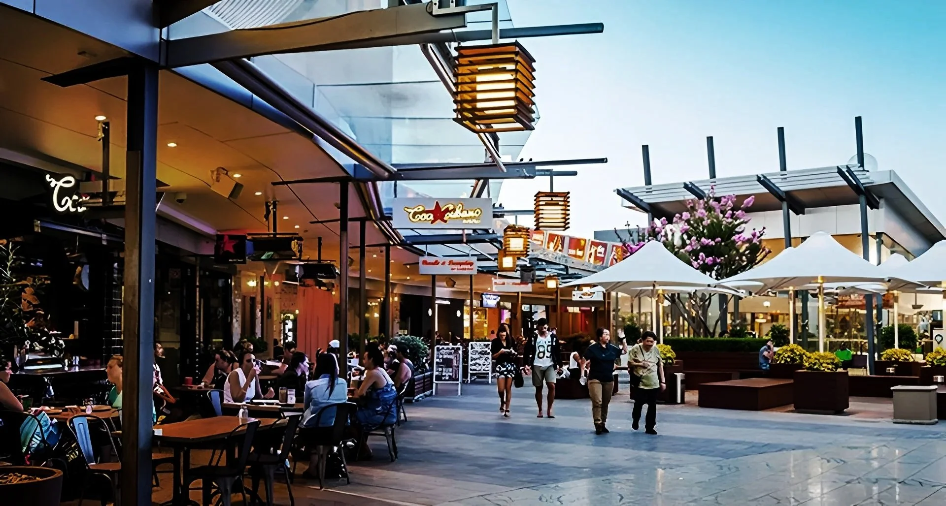 Outdoor shopping plaza with restaurants and cafes, people dining and walking, white umbrellas, pink flowering tree, and modern building architecture during evening.