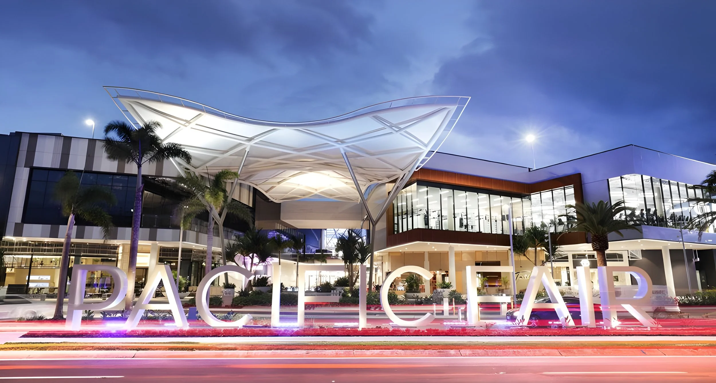 Night view of the Dadeland Mall in Miami, Florida, featuring a modern building with large glass windows, decorative tropical trees, and illuminated sign spelling 'DADELAND' in front.