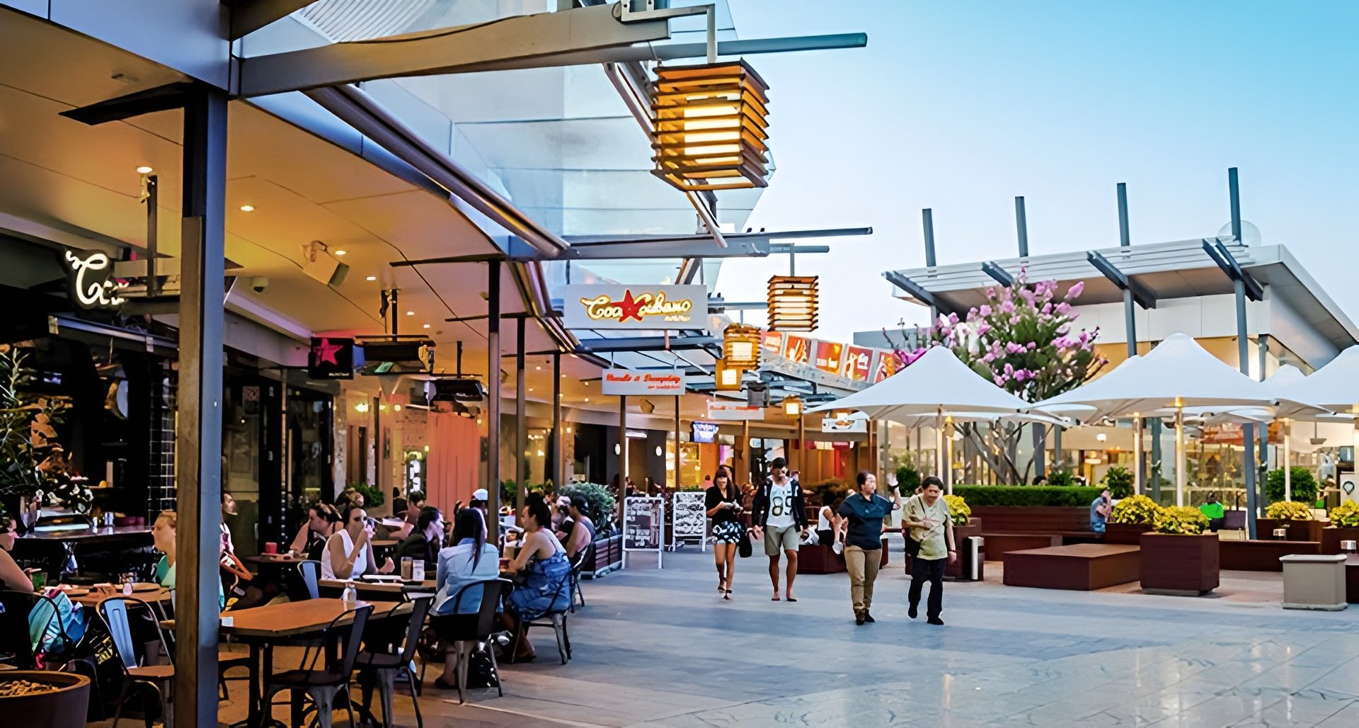 People dining and walking at an outdoor shopping and dining area with signs, trees, and white umbrellas.