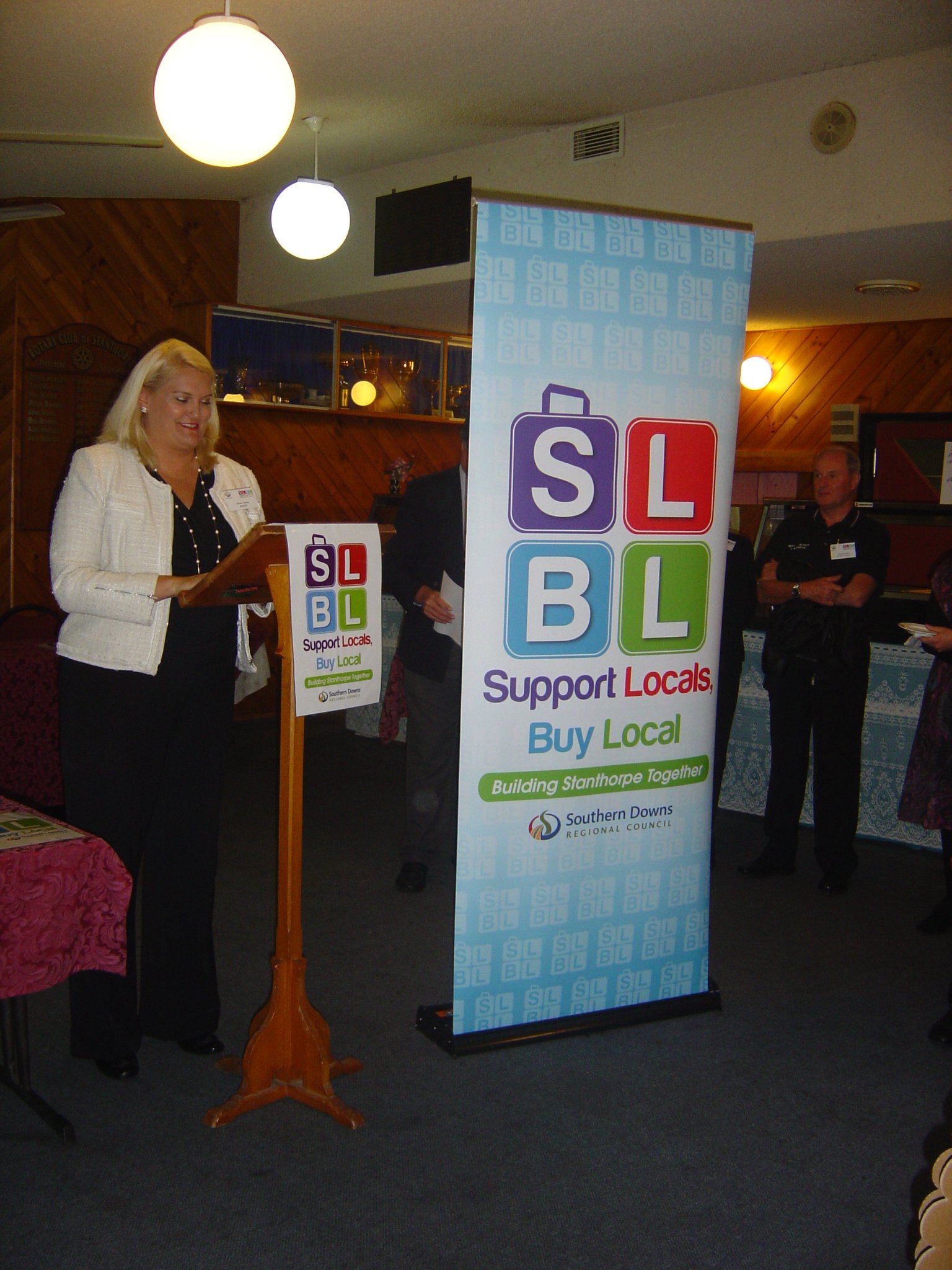 A woman standing at a podium reading a document during a presentation at an event supporting local businesses, with a large vertical banner displaying the SLBL logo and tagline Support Locals, Buy Local.