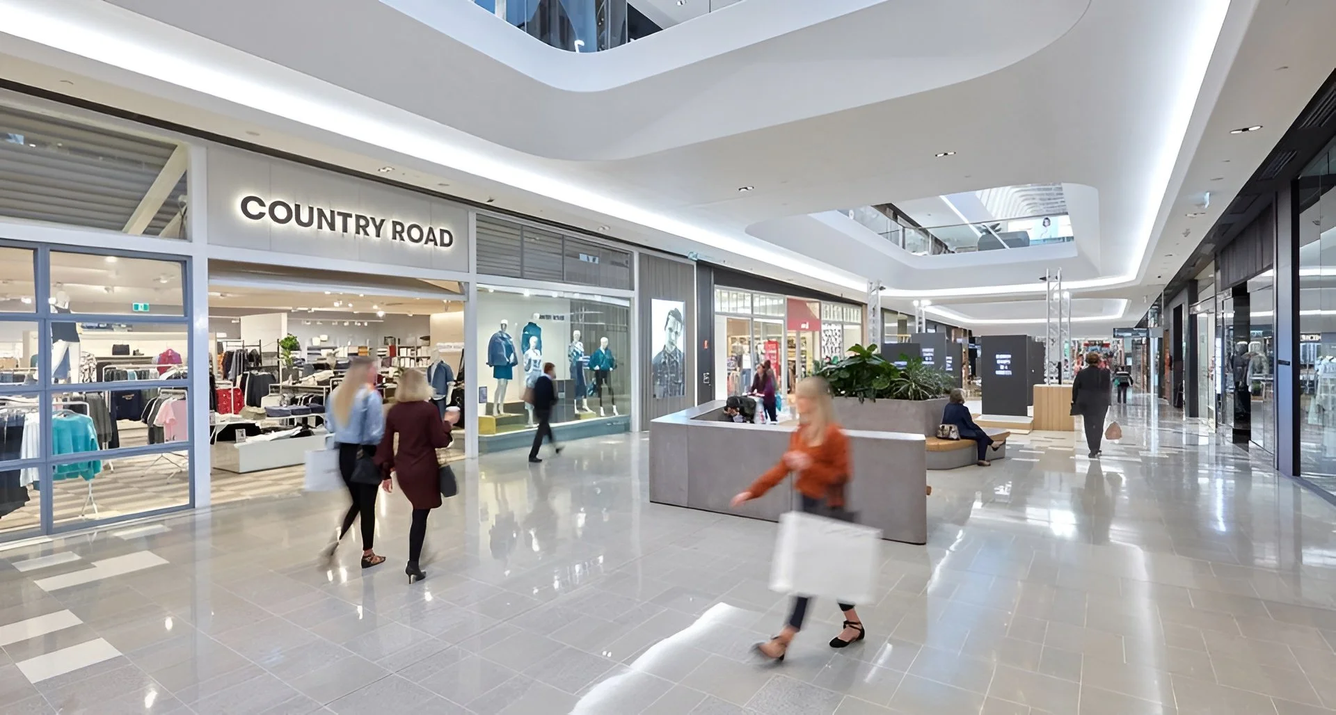 Interior of a shopping mall with customers walking near storefronts, including a clothing store named 'Country Road' and people sitting on benches, with bright lighting and a modern design.