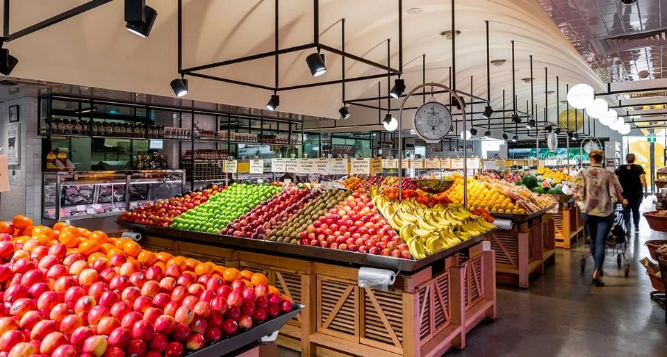 Inside a grocery store with fresh fruits on display, including apples, bananas, and other produce, with shoppers shopping.