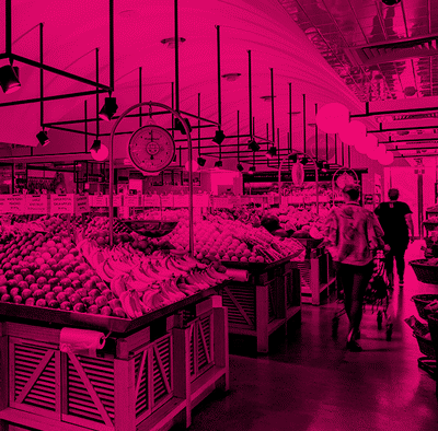 People shopping in a grocery store produce section with displays of fruits and vegetables, under modern lighting.