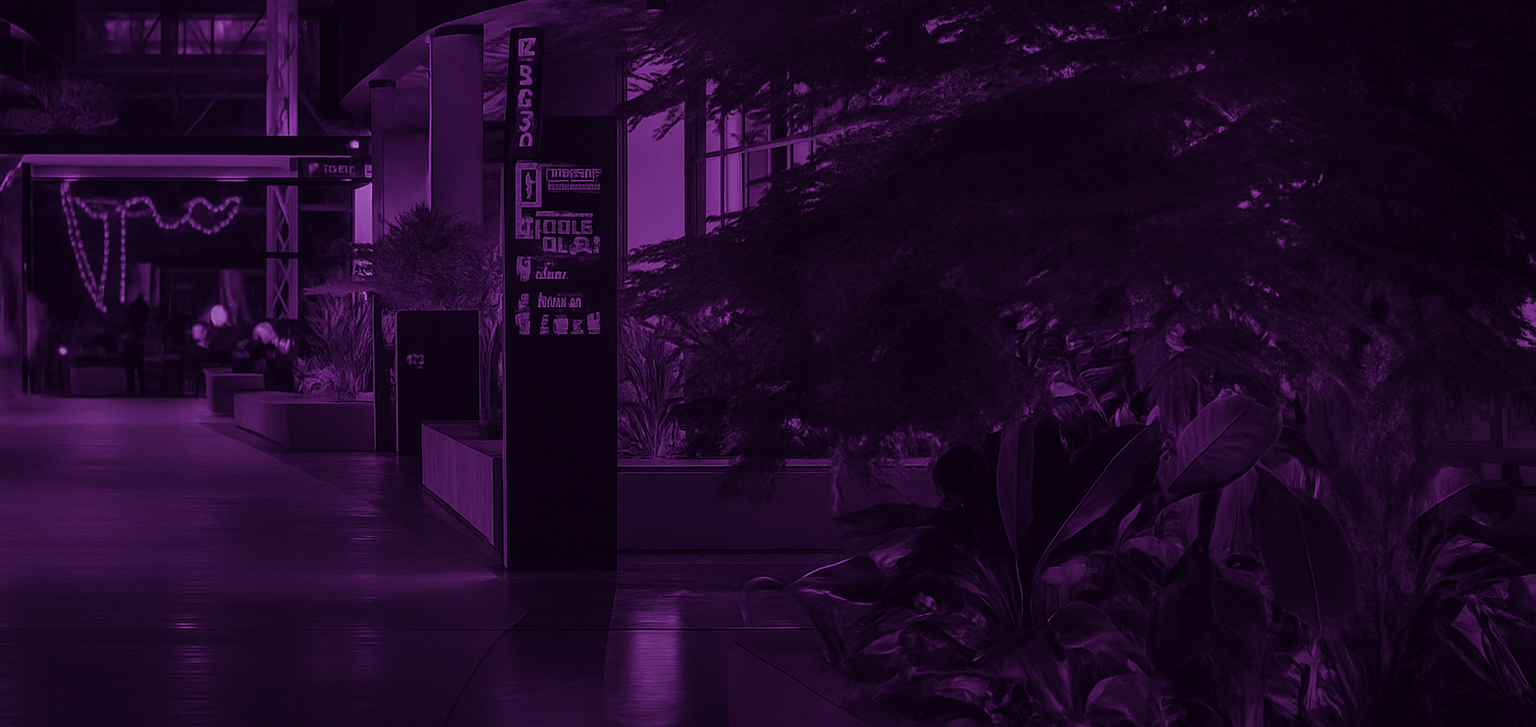 Night view of an indoor space illuminated with purple lighting, featuring large plants and an open walkway with benches and illuminated signs.
