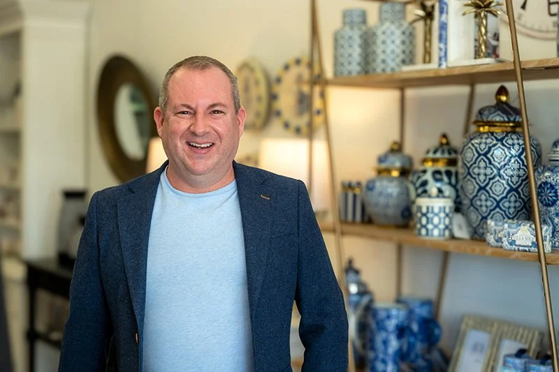Man smiling in a home decor store with blue and white ceramic vases on wooden shelves behind him.