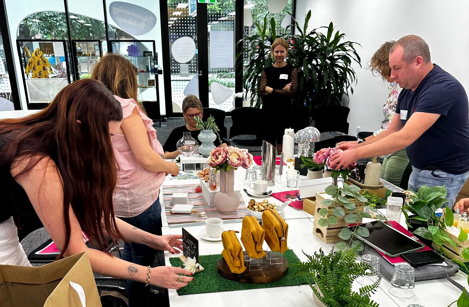 People gathered around a table with floral arrangements, cups, and various decorative items in a bright, modern indoor space, participating in a workshop.