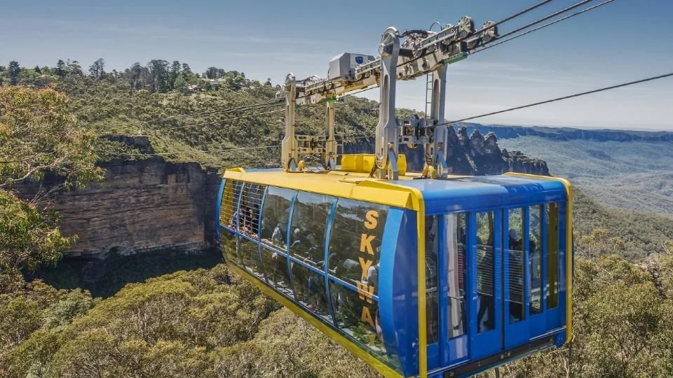 A blue and yellow sky tram cable car traveling above a lush green landscape with cliffs and hills in the background.